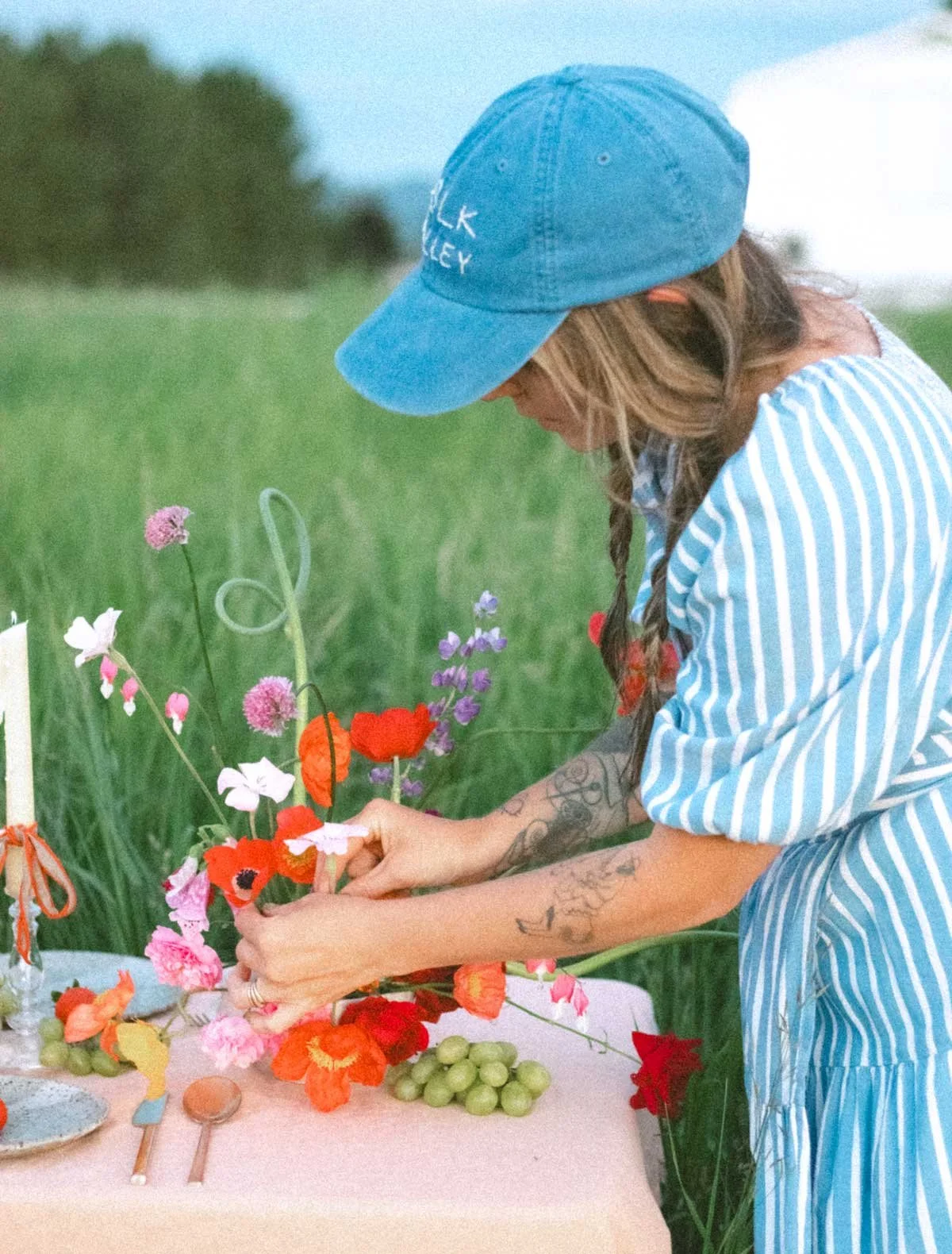 A woman in a striped dress and blue cap arranging flowers on a table outdoors.