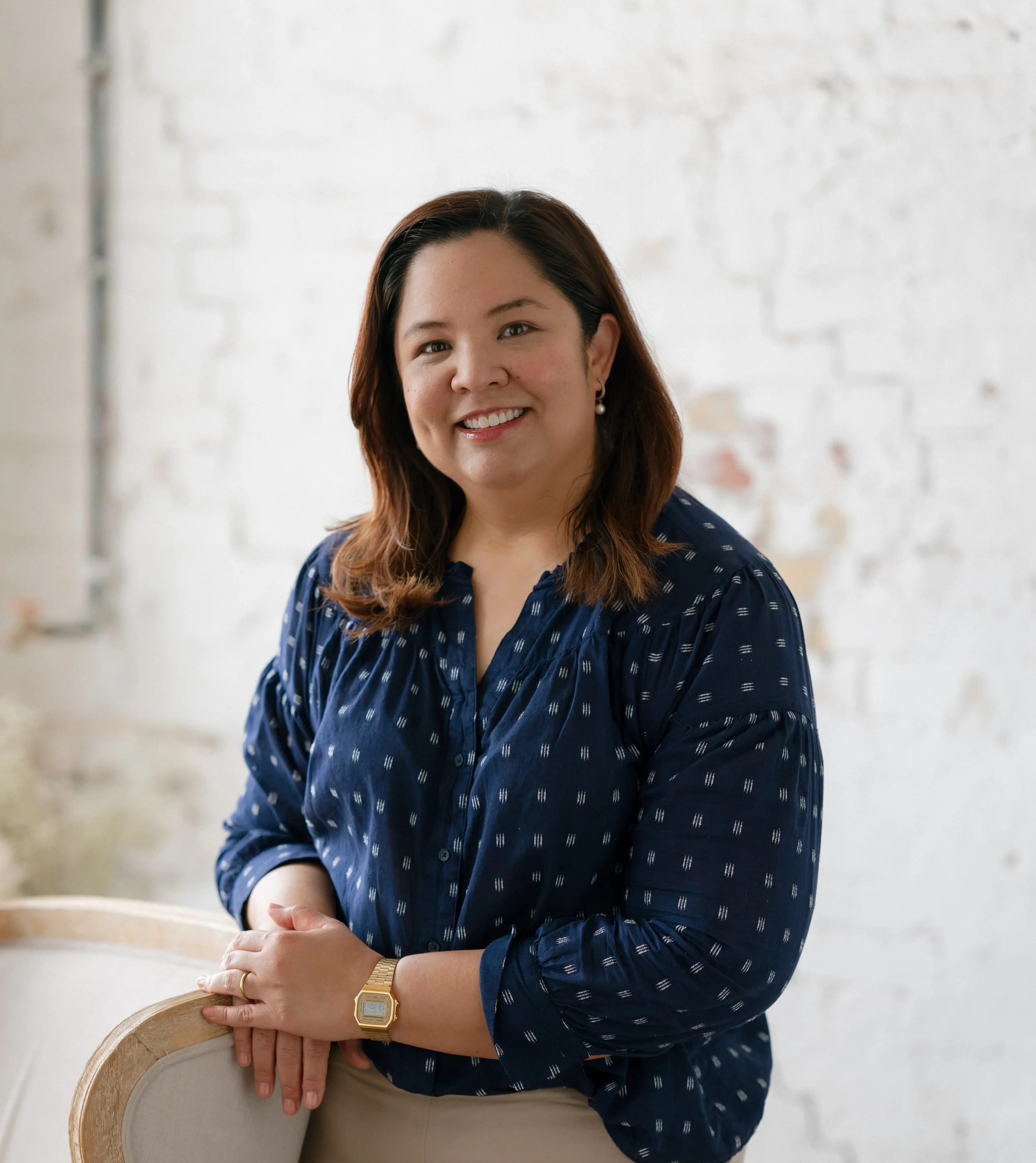 A woman with shoulder-length brown hair and earrings, wearing a dark blue patterned blouse, smiling and leaning on a wooden chair.