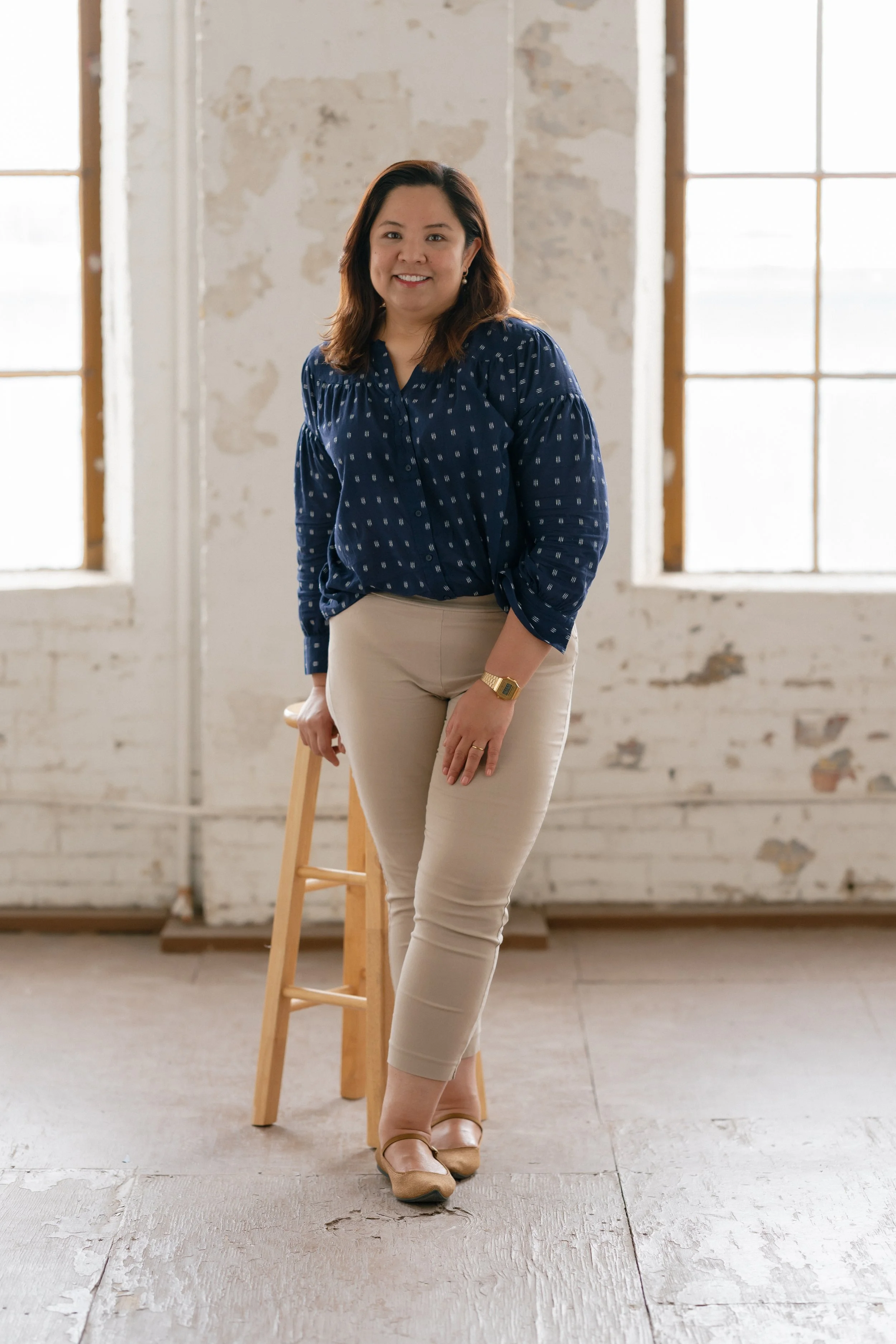 A smiling woman with shoulder-length dark hair standing in a bright, industrial-style room with large windows and white brick walls. She is dressed in a navy blue patterned blouse and beige pants, with a gold watch and tan shoes. She is holding onto a wooden stool.