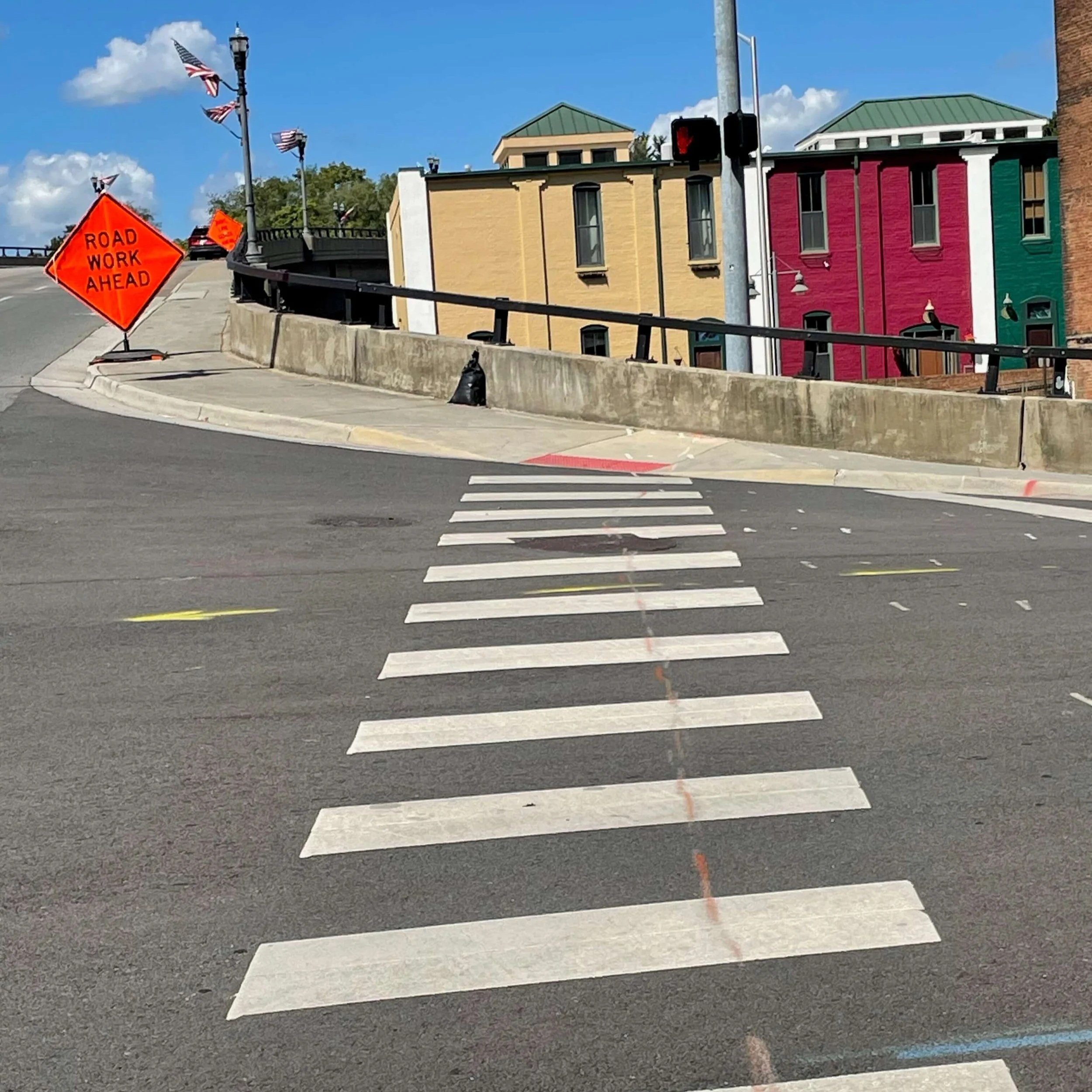 Street scene with a crosswalk in the foreground, colourful buildings in yellow, pink, green, and red, an orange road work sign, and a traffic light showing red, under a blue sky with some clouds.
