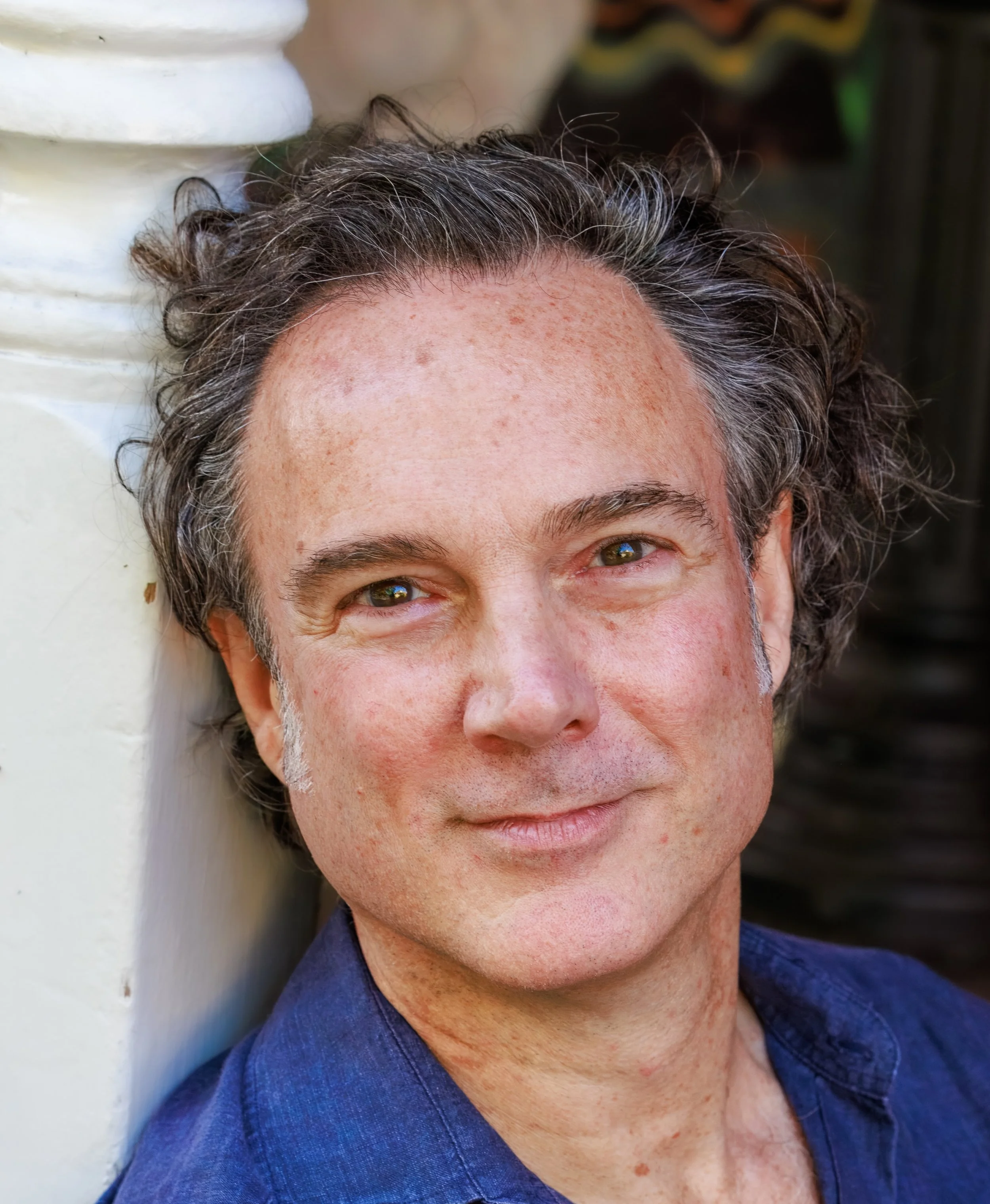 Close-up of a smiling middle-aged man with curly gray hair, wearing a blue shirt, outdoors near a white column with a dark background.