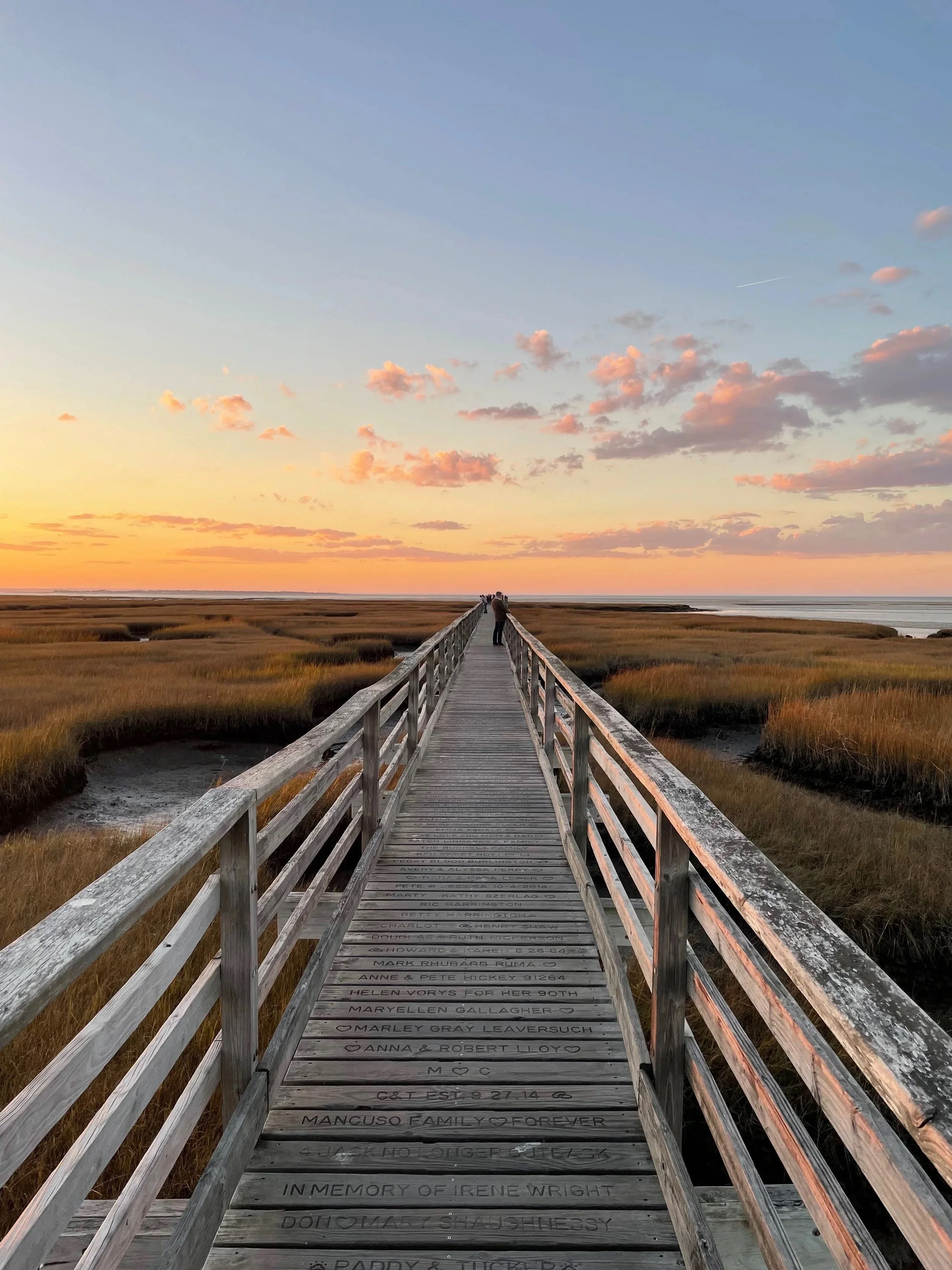 A long wooden boardwalk extending over marshy grassland during sunset, with dozens of engraved messages and names on the planks, and a few people visible in the distance at the horizon.