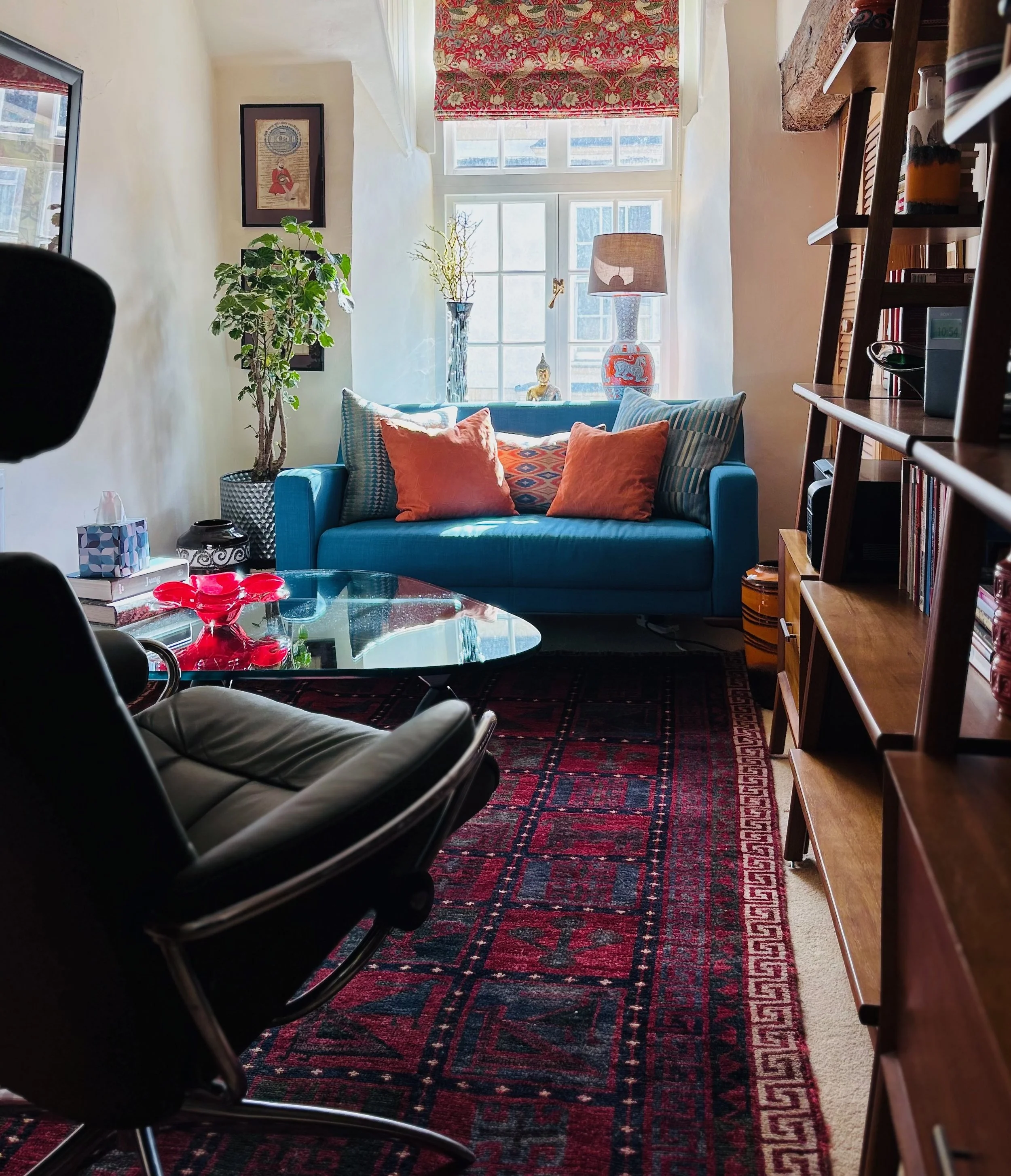 Therapy room with a large window, blue sofa with orange and patterned pillows, a tall planter with a leafy plant, a glass coffee table with red decorative bowl, a wooden bookshelf, and a patterned red and blue rug.