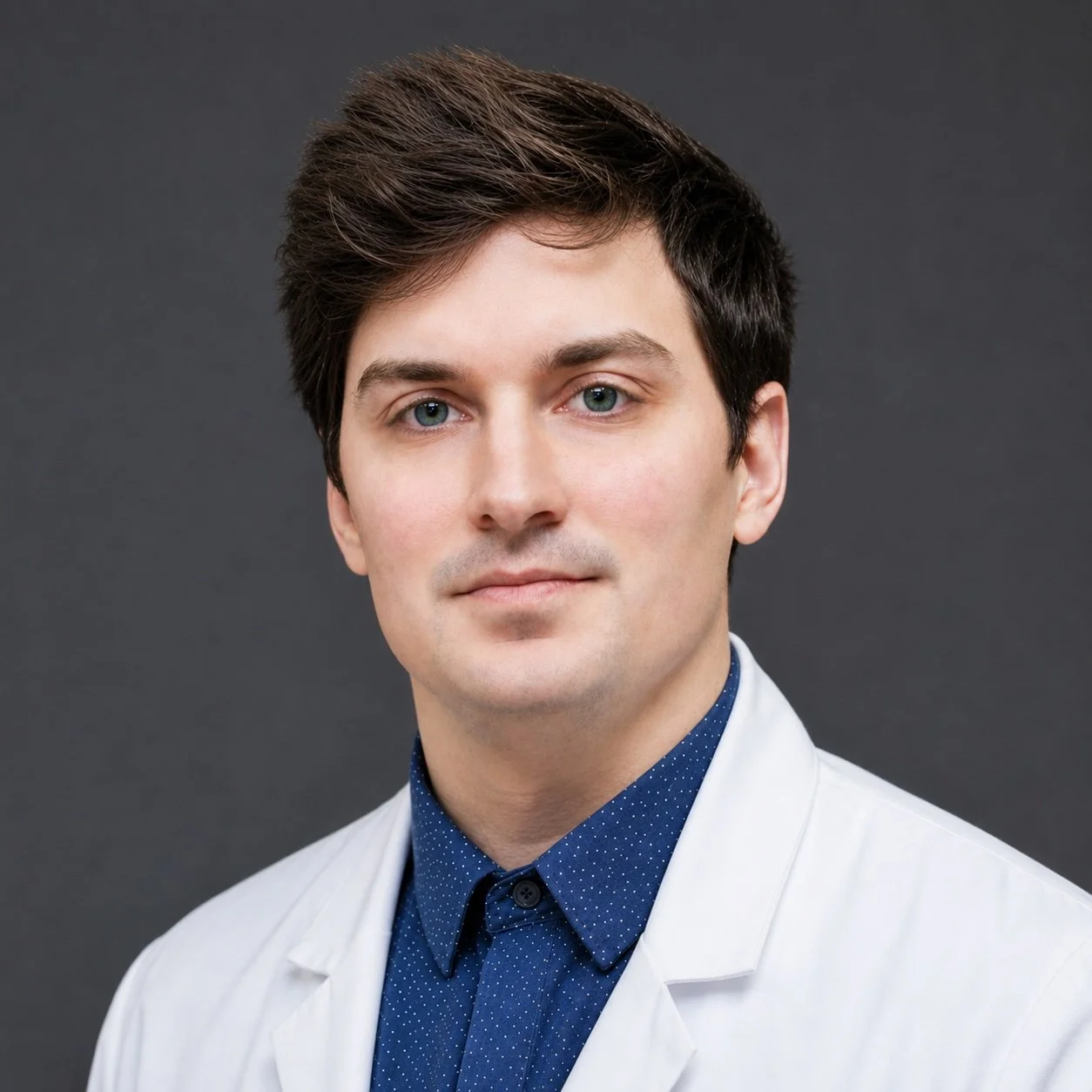 A young man with dark brown hair, blue eyes, and light skin, wearing a white medical coat and a blue dotted shirt, standing against a dark gray background.
