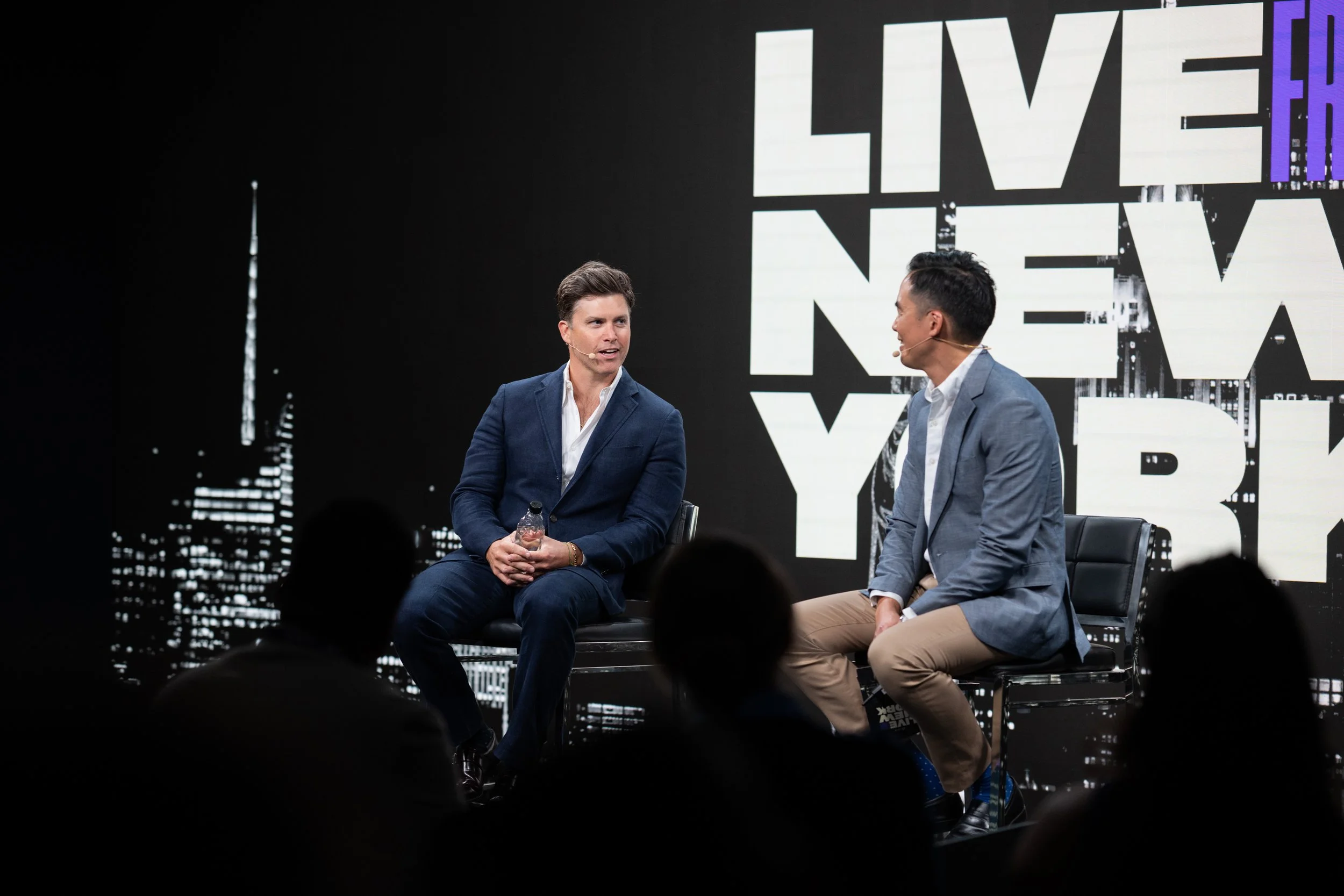 Two men sitting on stage during a panel discussion with a city skyline background and the word 'LIVE' in large letters behind them.