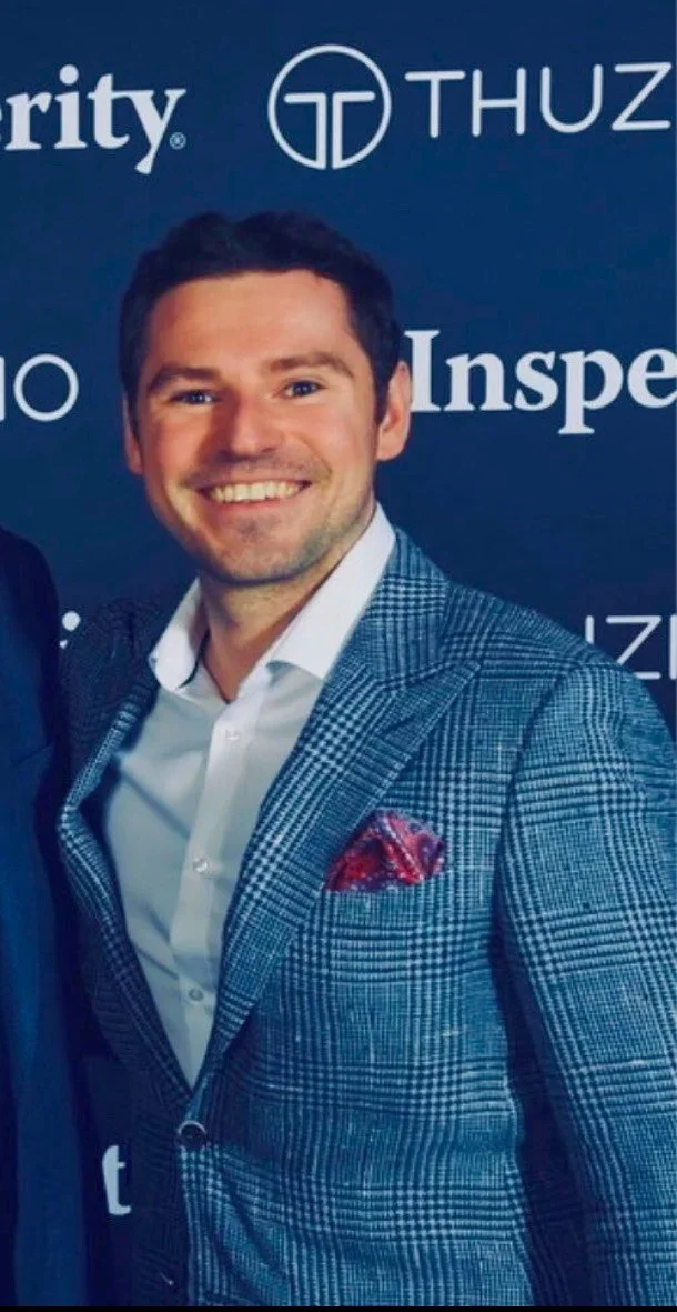 A smiling man in a blue patterned suit and white shirt, standing in front of a dark backdrop with logos and text.