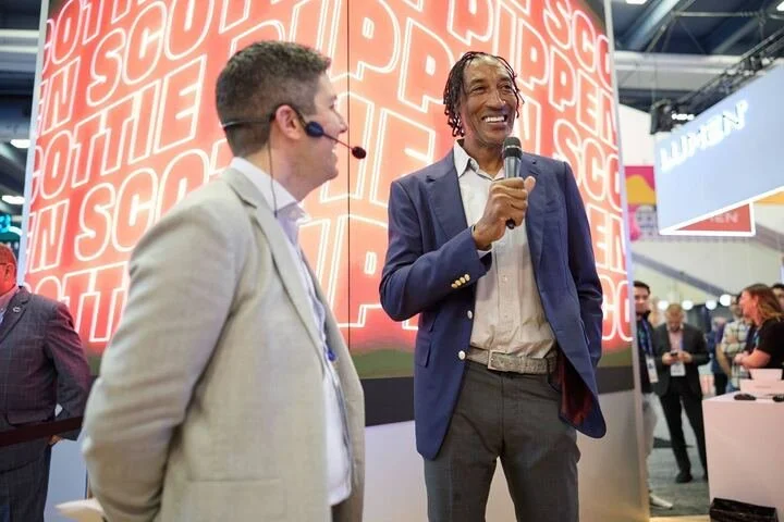 Two men speak at a conference or event, with a large red background reading 'SCOP' behind them. The man on the left wears a light-colored blazer and headset, while the man on the right, holding a microphone, wears a dark blue blazer and gray trousers.