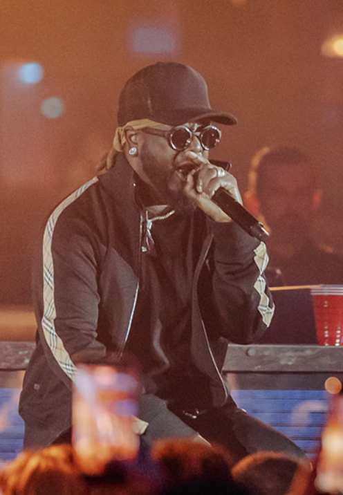 A man performing on stage, wearing sunglasses, a cap, and a black jacket with white stripes, holding a microphone, with a blurred crowd in the foreground.