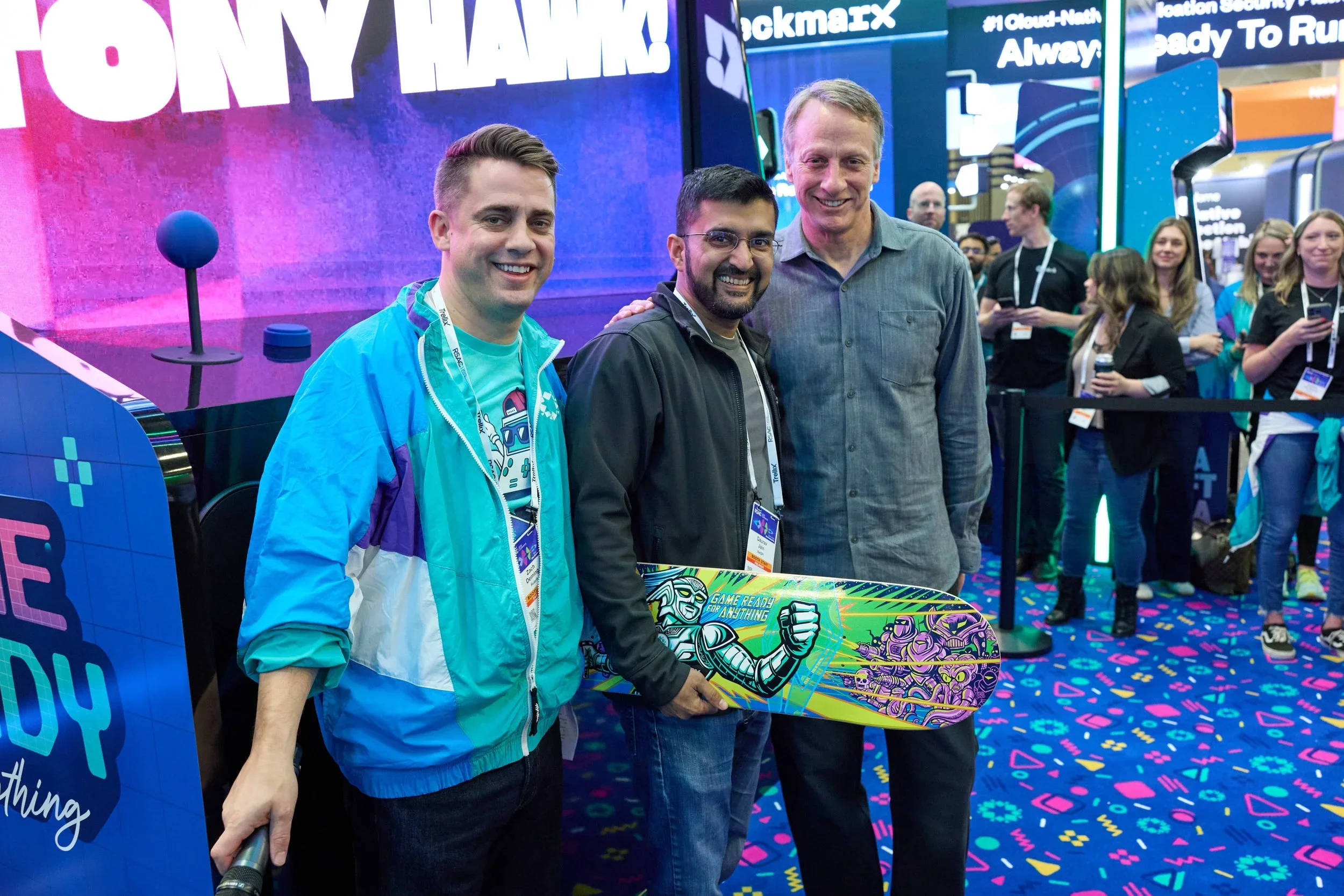 Three men standing together at a trade show or convention, smiling at the camera. The background features colorful electronic displays and a crowd of people.