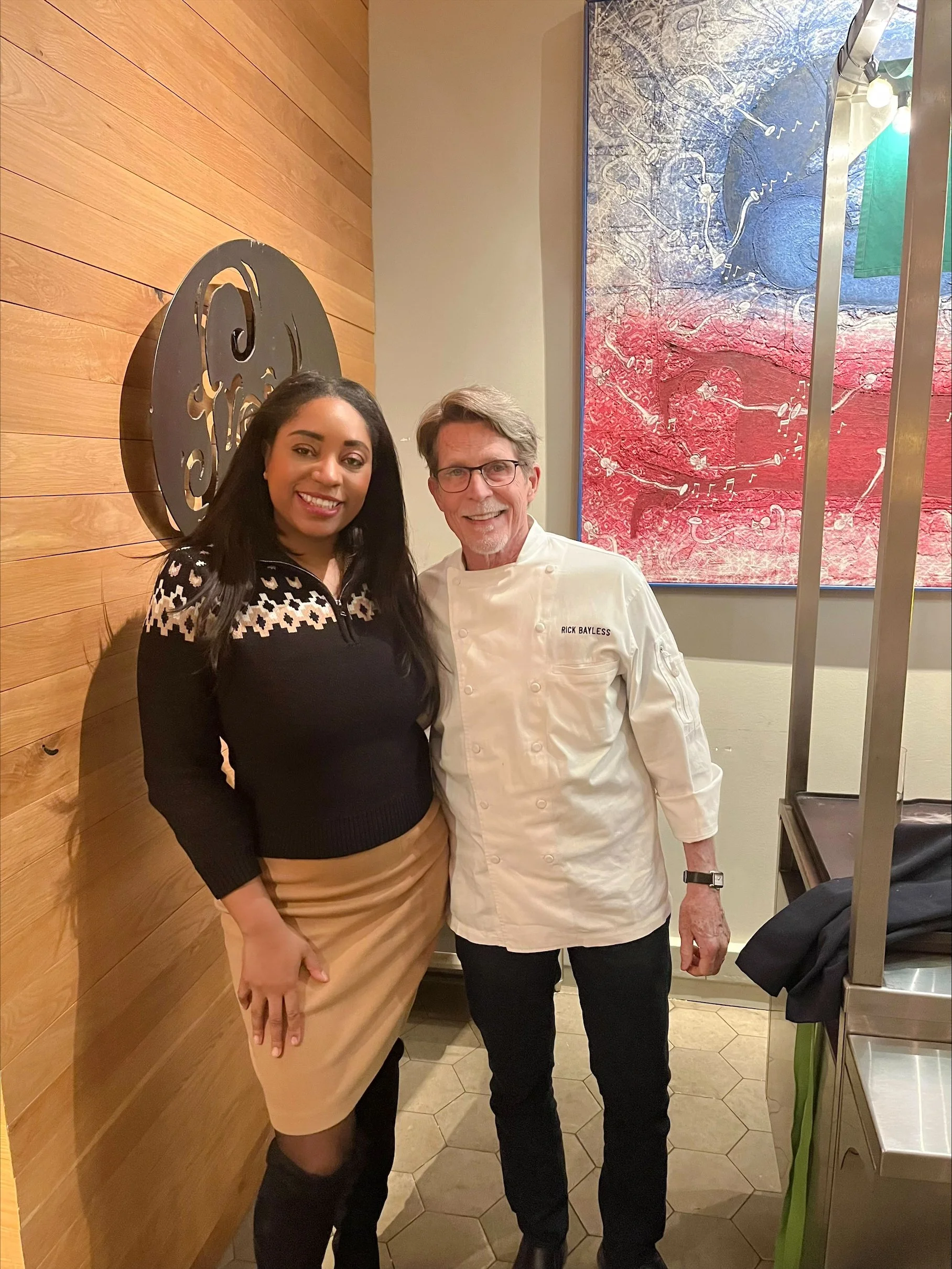 A woman and a man standing together in a restaurant. The woman is wearing a beige skirt, black stockings, and a black sweater with a white pattern. The man is dressed in a white chef's jacket with the name "Rick Bayless" embroidered on it and glasses. They are smiling and posing for the photo.