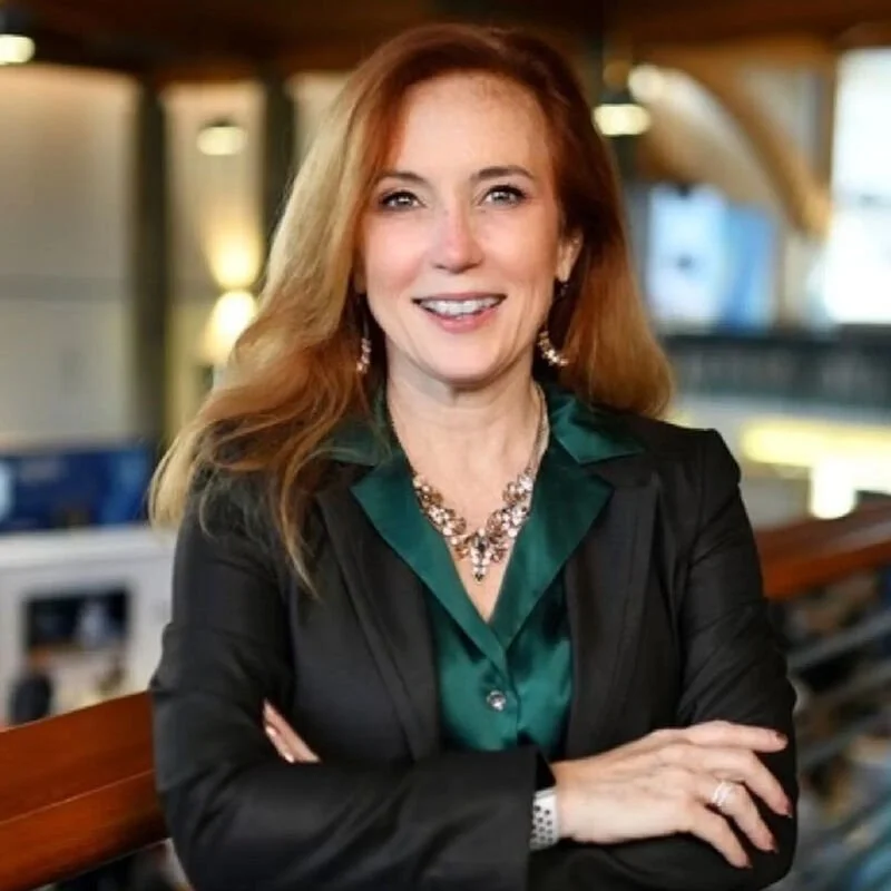 A woman with red hair wearing a black blazer and green blouse, smiling with arms crossed, in an indoor setting with warm lighting.