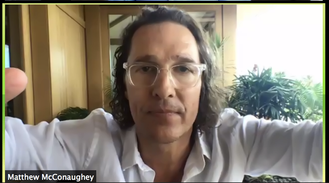 A man with curly hair, glasses, and wearing a white shirt, is taking a selfie in a room with wooden paneling and abundant greenery in the background.