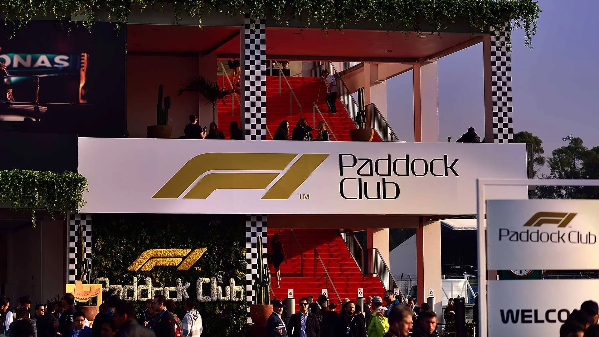 Entrance to the Paddock Club with a large sign displaying the F1 logo and text, a red staircase, and people gathered outside during daytime.