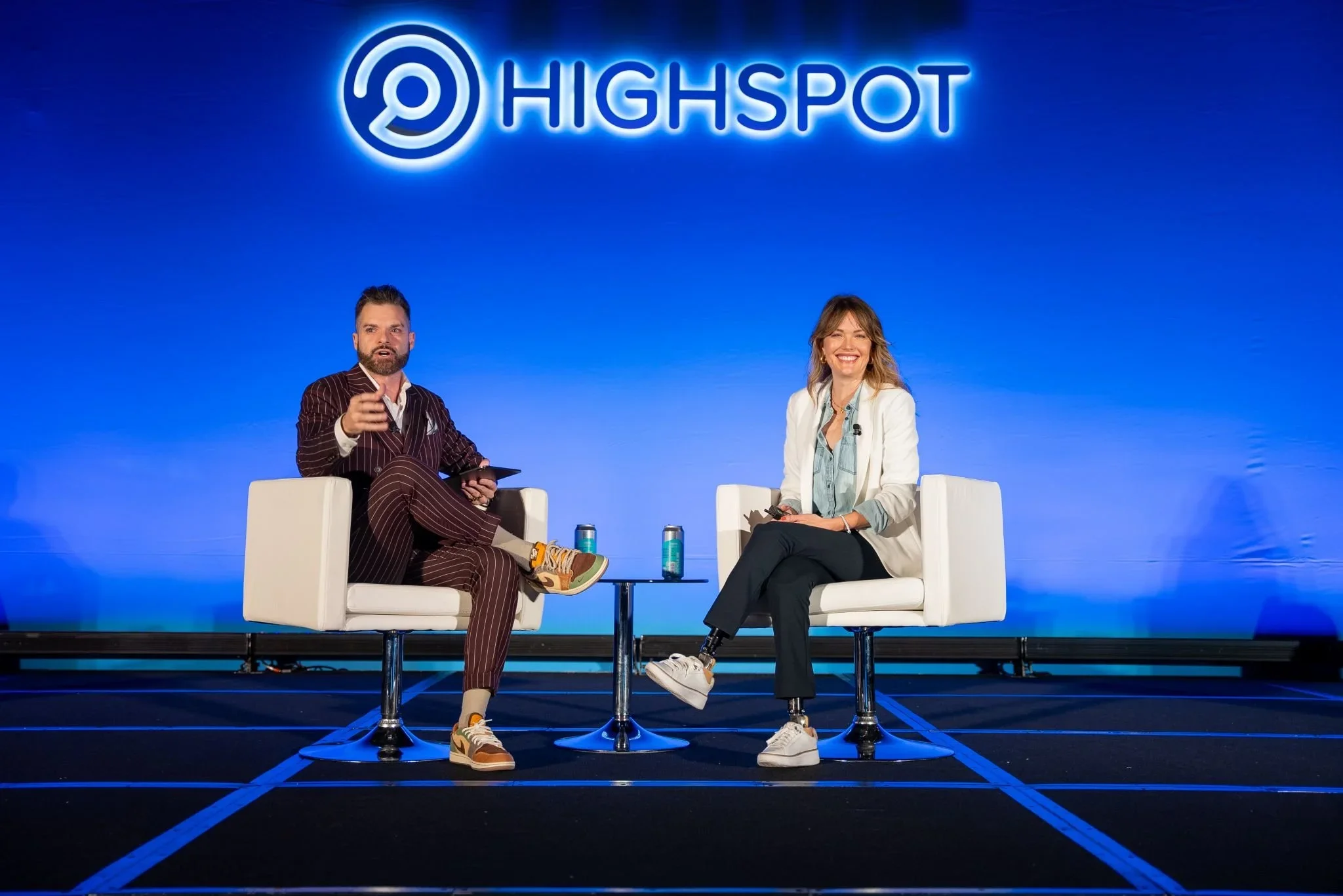 Two people sitting on stage under a blue neon sign that reads 'Highspot' with a logo; man on the left in a striped suit and sneakers, woman on the right in a white blazer and casual attire, both smiling and holding microphones.