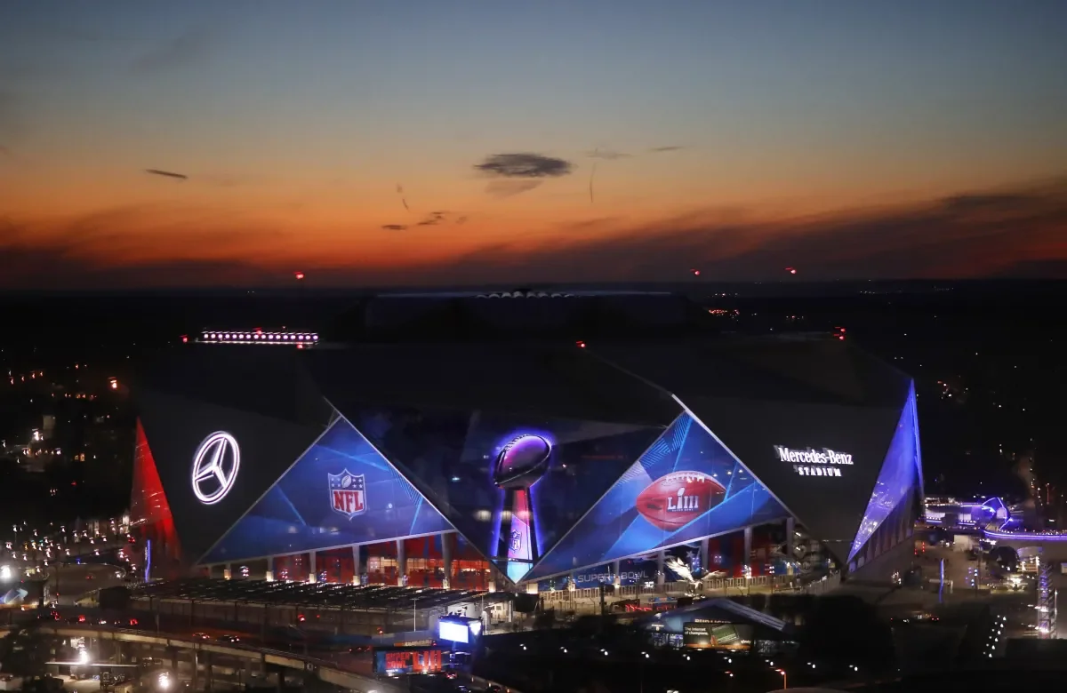 Nighttime view of a large, modern football stadium decorated with Super Bowl logos, Mercedes-Benz branding, NFL emblem, and flags, illuminated against a dark sky with a colorful sunset or sunrise in the background.