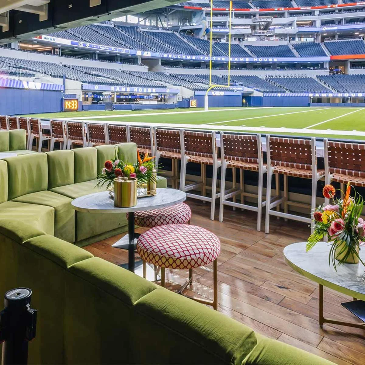 Luxury lounge view inside a sports stadium, with green seating, floral arrangements on marble tables, and empty stadium seats and field in the background.