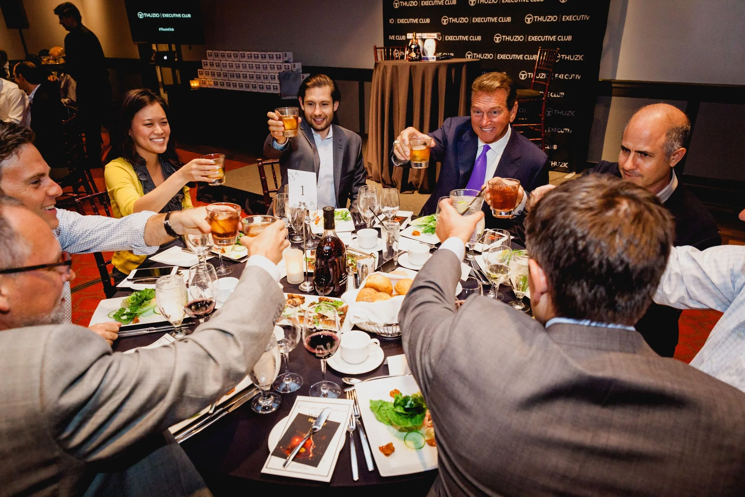 A group of diverse people in formal attire sitting around a table at a celebratory dinner event, raising their glasses in a toast, with plates of food and drinks on the table. In the background, a man in black and a large screen display the event branding for 'Thuzio Executive Club'.