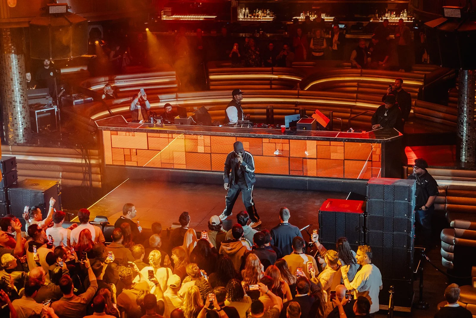 A performer on stage at a nightclub with a large crowd watching. The stage has a DJ setup, and the performer is dressed in black with a hoodie, speaking into a microphone. Several people are recording the event with their phones, and there are people in the background near the bar area.