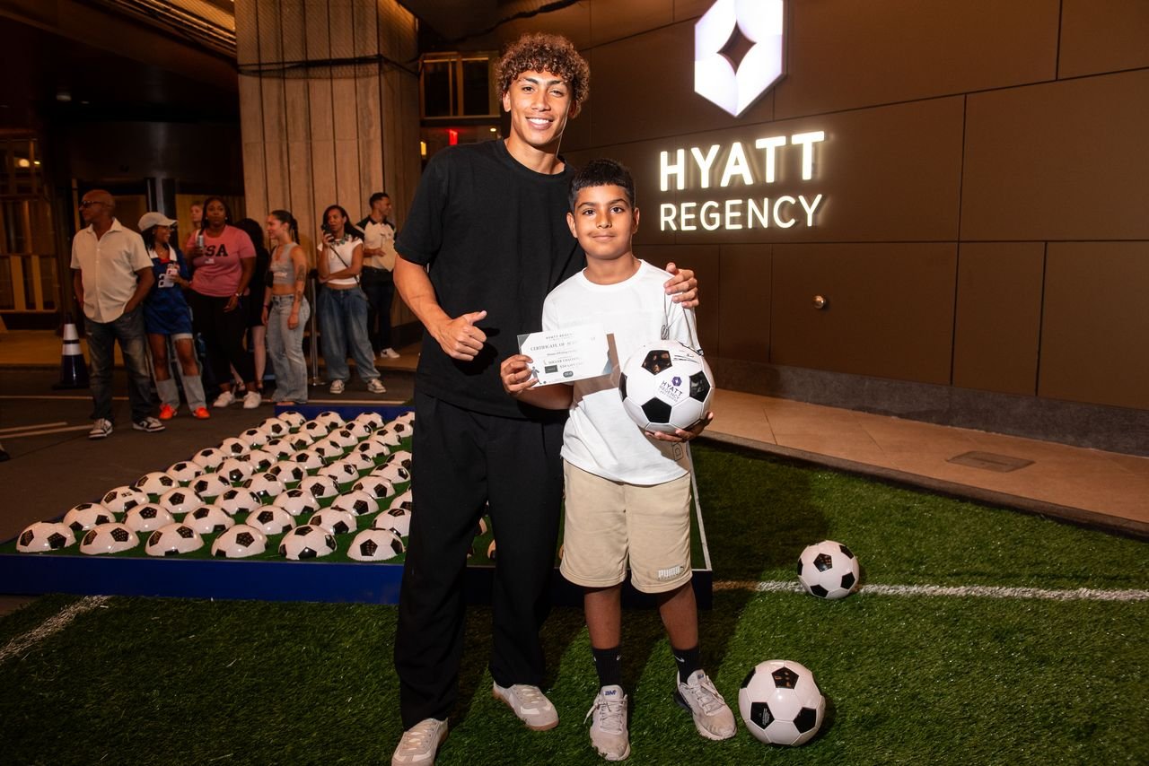 A young man and boy standing outside the Hyatt Regency hotel at night. The boy is holding a soccer ball and an envelope, with more soccer balls around their feet. In the background, people are lined up near a display of many soccer balls.
