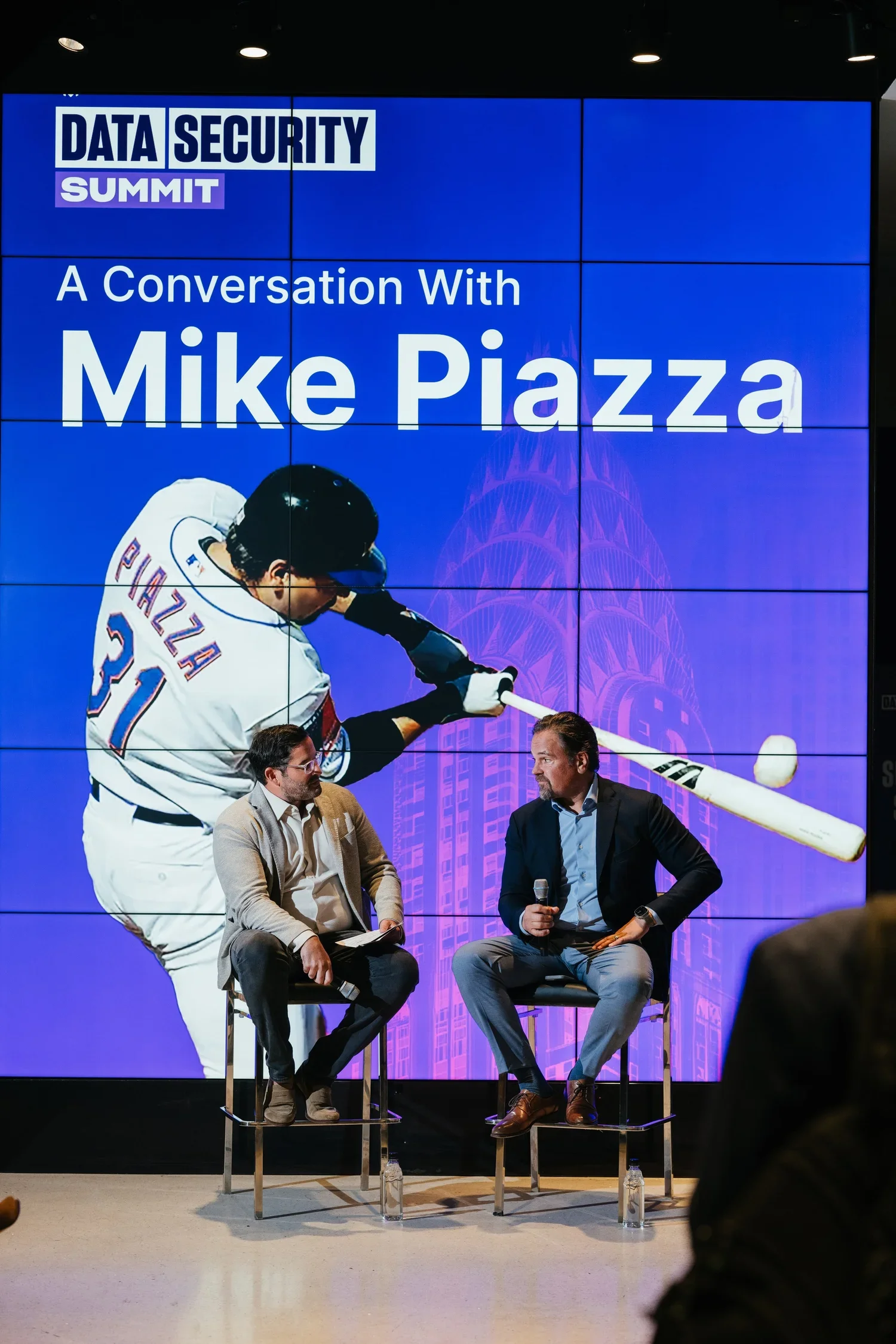 A panel discussion at a data security summit featuring a large screen with the summit's title. Two men are seated on stools in conversation; one holds a microphone.