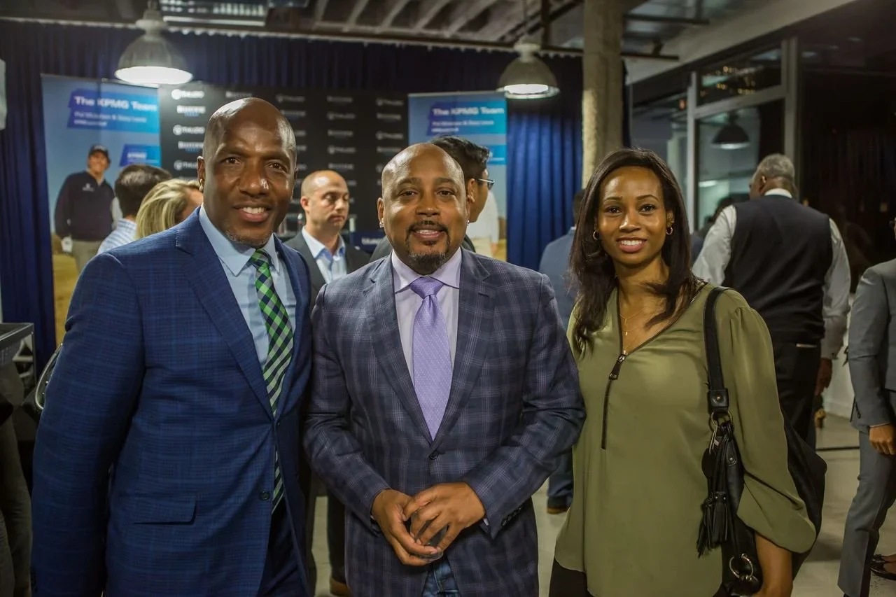 Three professionally dressed people smiling at the camera at an indoor event or conference, with other attendees in the background.