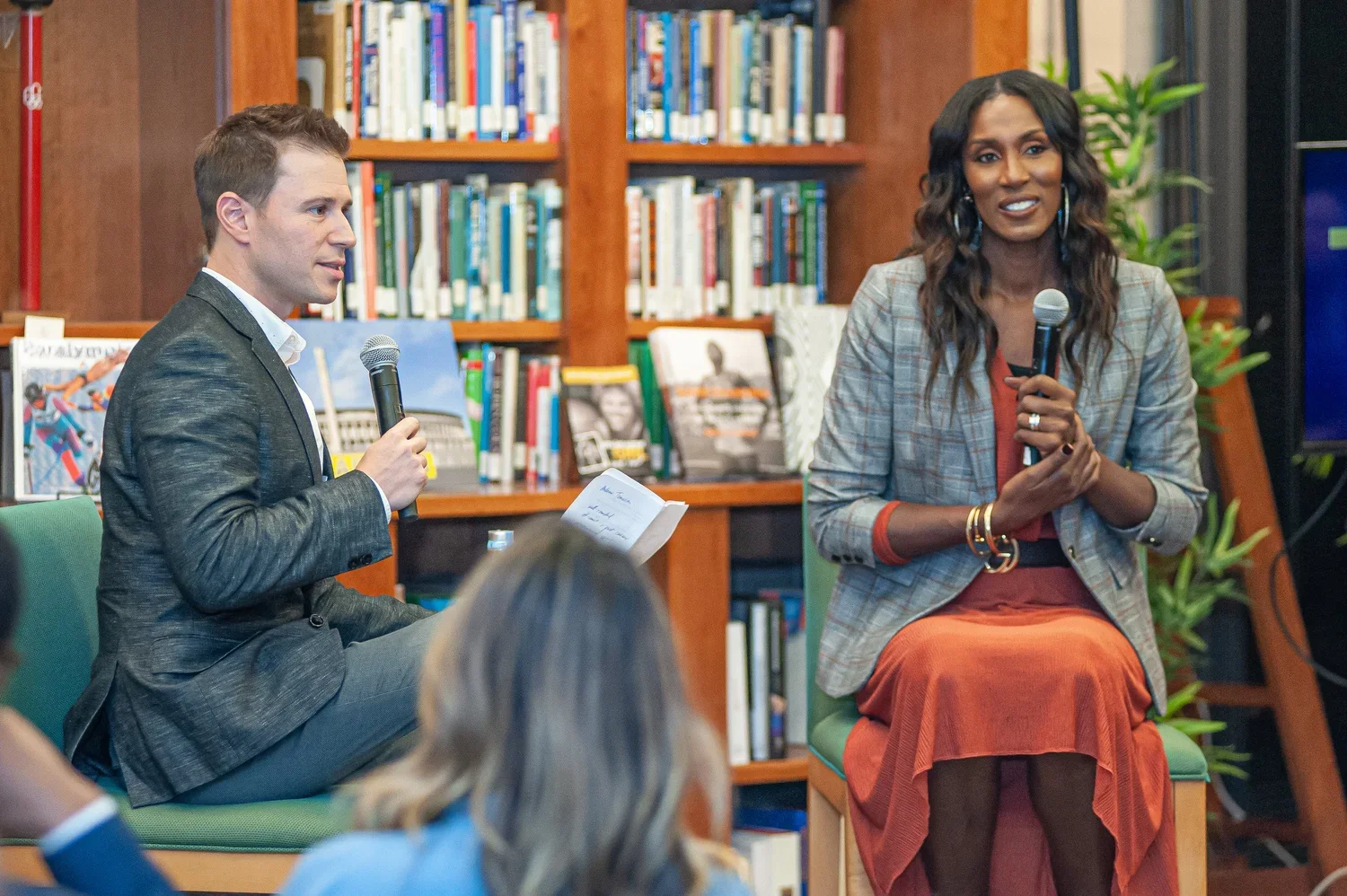 Two people sitting on chairs, holding microphones, engaging in a discussion in a library or bookstore setting with bookshelves in the background.