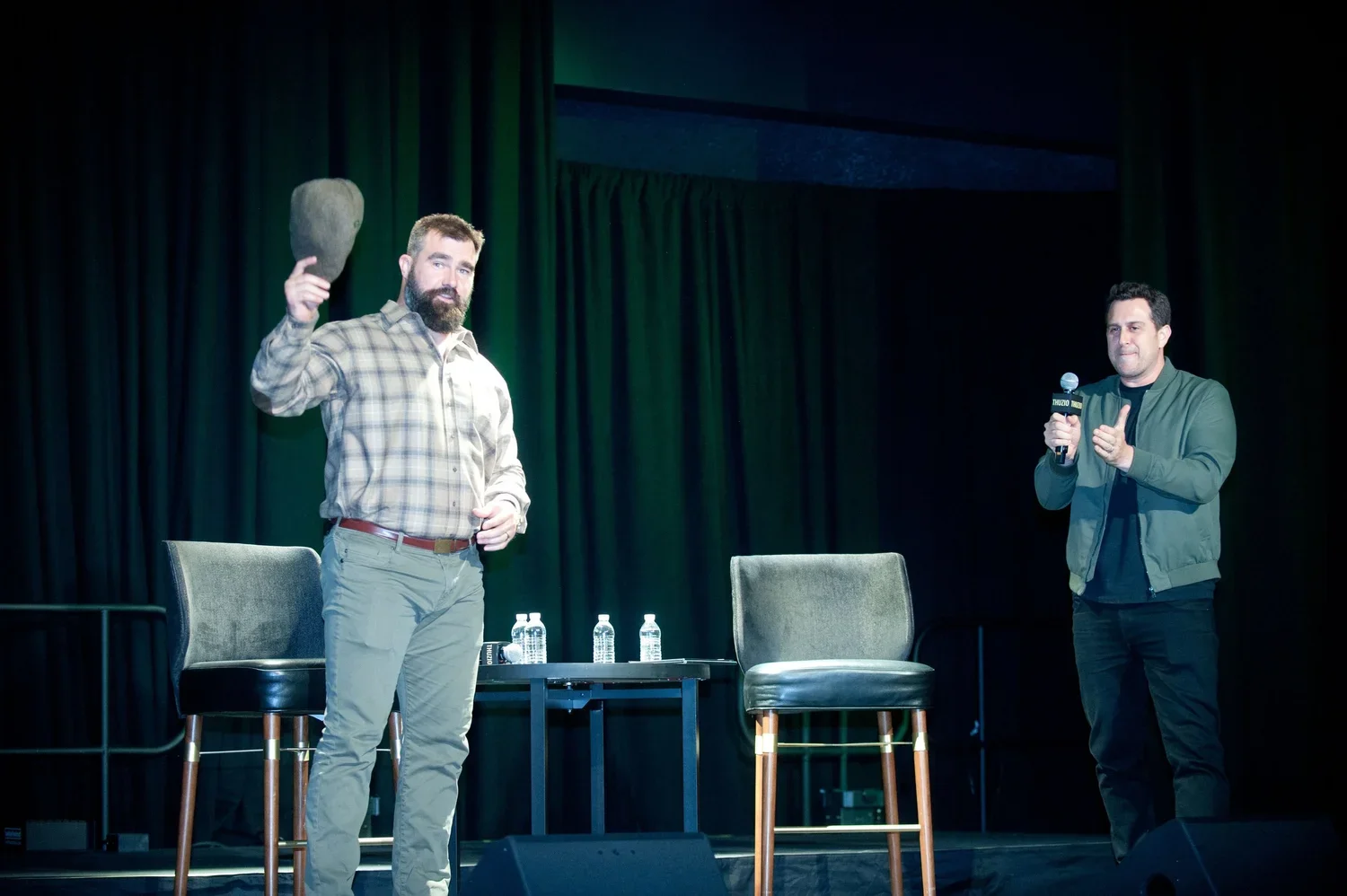 Two men on stage during a panel discussion or talk. One man on the left is holding a rock and has a beard, wearing a plaid shirt and khaki pants. The other man on the right is holding a microphone, wearing a dark jacket and black pants. There are two empty chairs, a small table with water bottles between the men, and a black curtain backdrop.