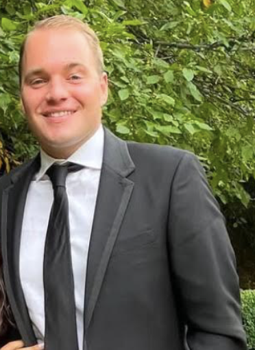 A smiling young man in a black suit, white shirt, and black tie standing outdoors with green foliage in the background.