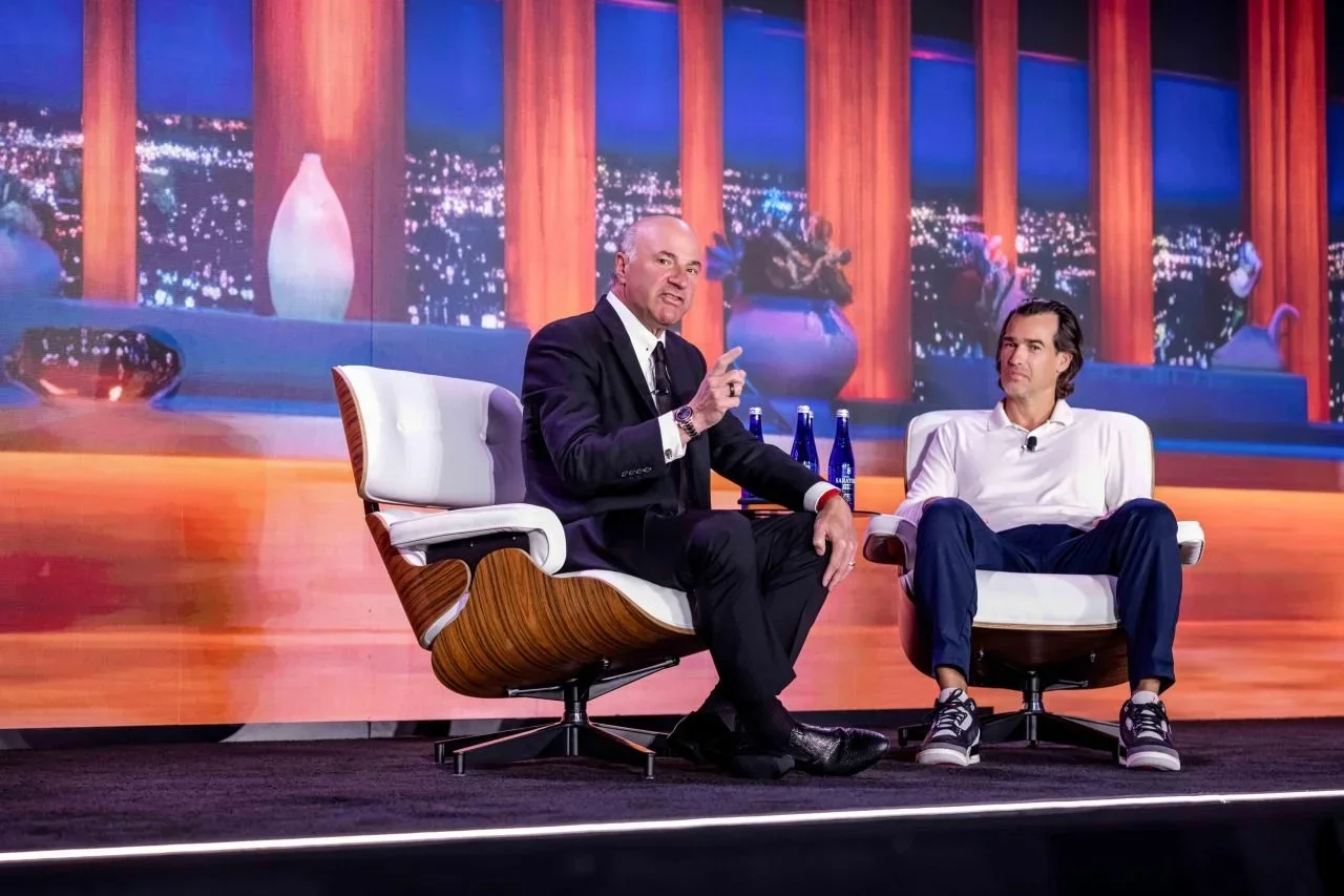 Two men sitting on stage chairs in a TV studio with a backdrop of a city skyline at night. One man is dressed in a black suit and white shirt, gesturing with his right hand. The other man is dressed in a white shirt and blue pants, sitting with legs apart. There are three bottles of water on a small table between them.