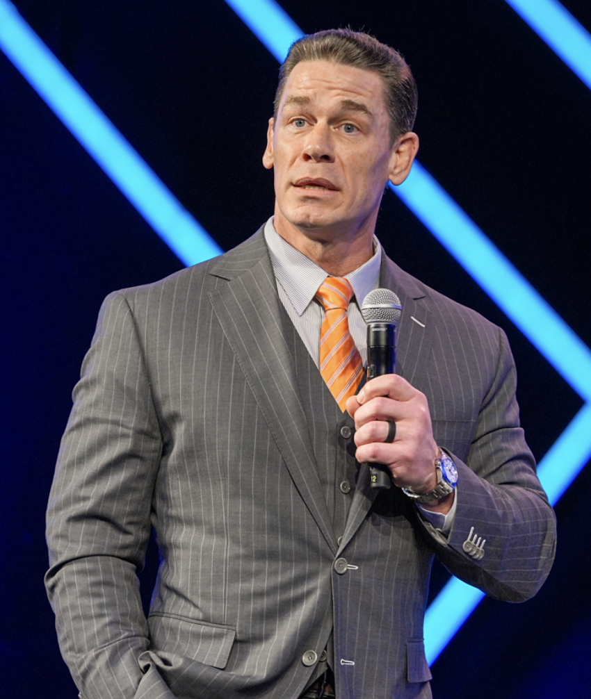 A man in a gray pinstripe suit and orange striped tie holding a microphone, standing on a stage with blue diagonal stage lights in the background.
