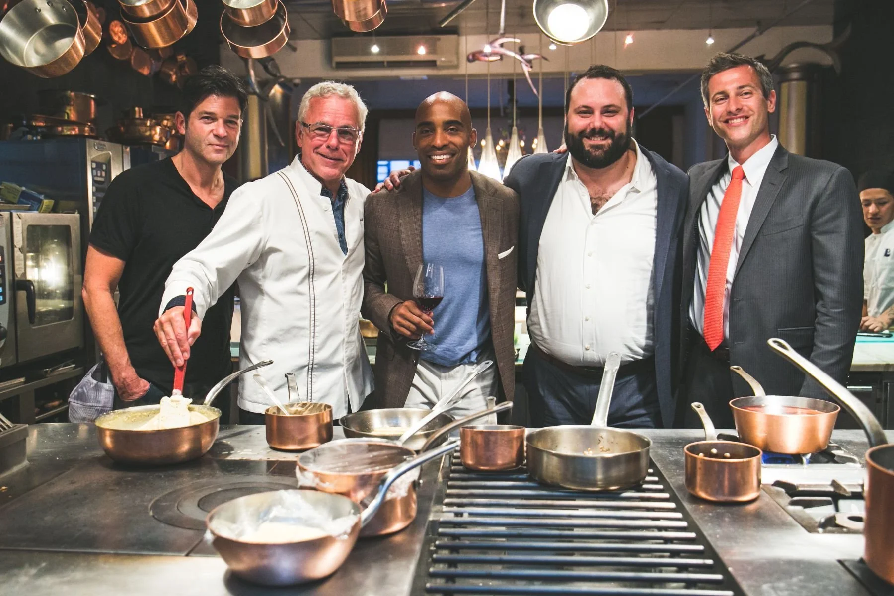 Group of five men smiling and posing for a photo in a professional kitchen with copper pots hanging from the ceiling. One man is holding a glass of red wine, and there are cooking utensils and pots on the counter in front of them.
