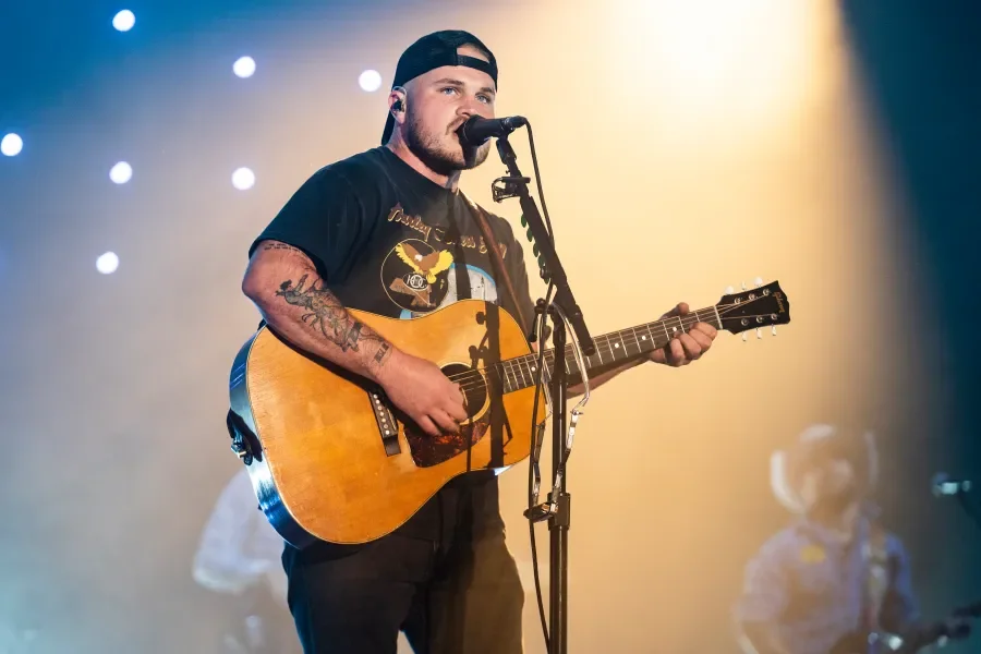 A man with tattoos wearing a black cap and t-shirt, playing an acoustic guitar and singing into a microphone on stage with stage lights in the background.