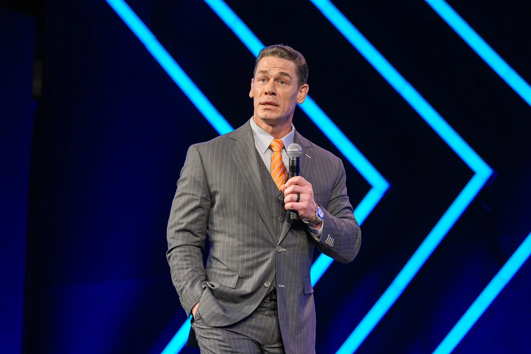 A man in a gray pinstripe suit, white shirt, and orange striped tie holding a microphone, speaking or presenting on stage with blue light design in the background.