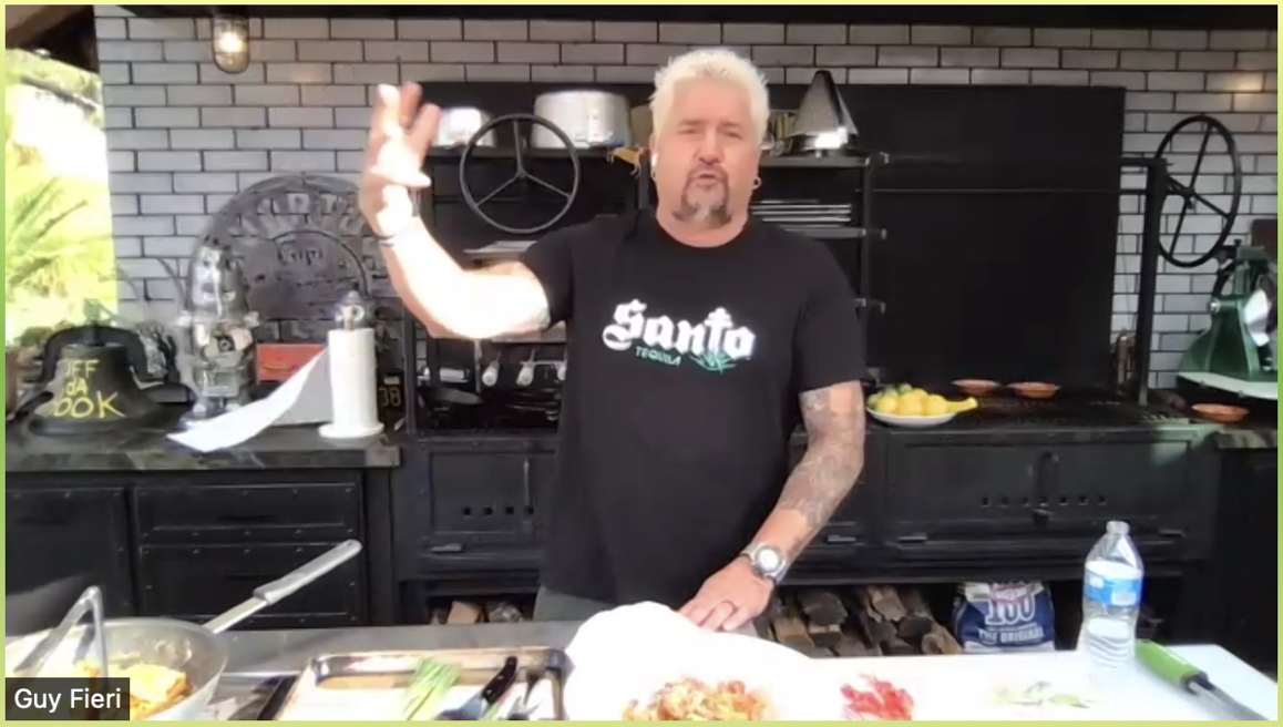 Man with platinum blonde hair and tattoos standing at a kitchen counter, making a hand gesture, in front of a grill and brick wall, with various kitchen items around.
