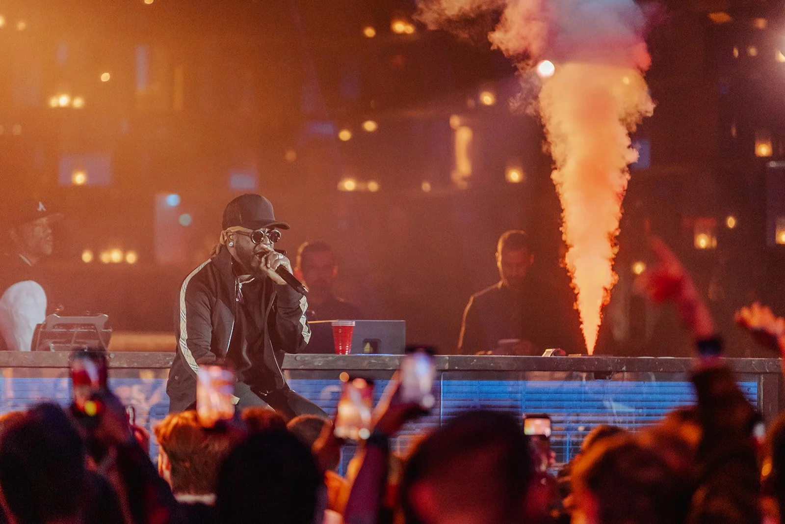 A performer wears sunglasses, a black cap, and a black jacket with white stripes on the sleeves, singing into a microphone on a stage at a concert. Behind him, an entertainer or DJ operates equipment, and a red smoke or fire eruption is visible on stage amidst dim lighting.