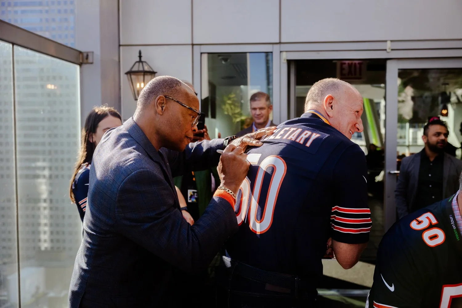 A man in a suit is signing a football jersey worn by another man, who is smiling. Several people are watching and taking photos, with a cityscape in the background.