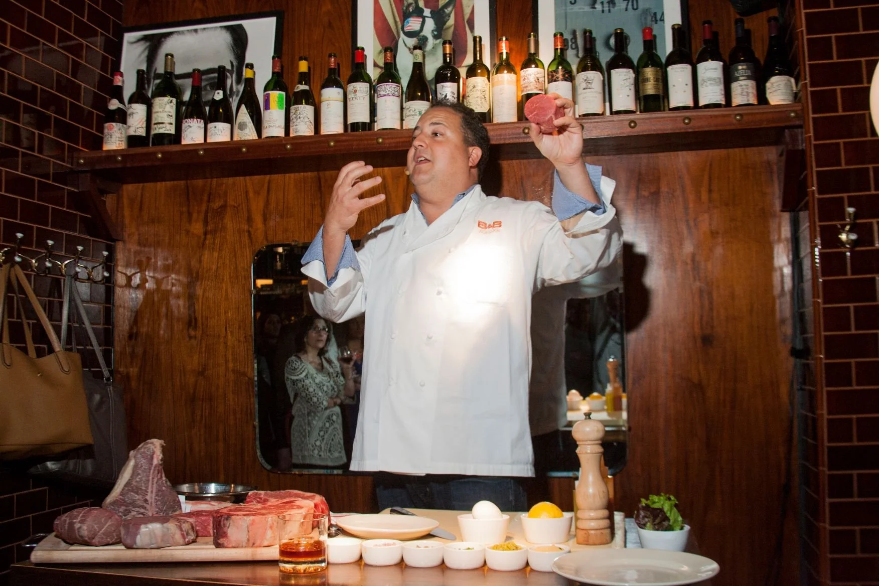 A man in a chef's coat holding a piece of raw meat, standing behind a table with various ingredients and spices, with a woman observing in the background.
