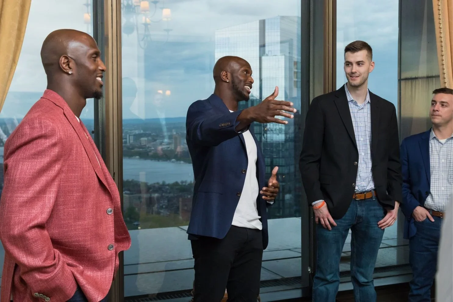 Four men in business attire having a conversation in a high-rise office with large windows overlooking a cityscape and water, some smiling and engaging.