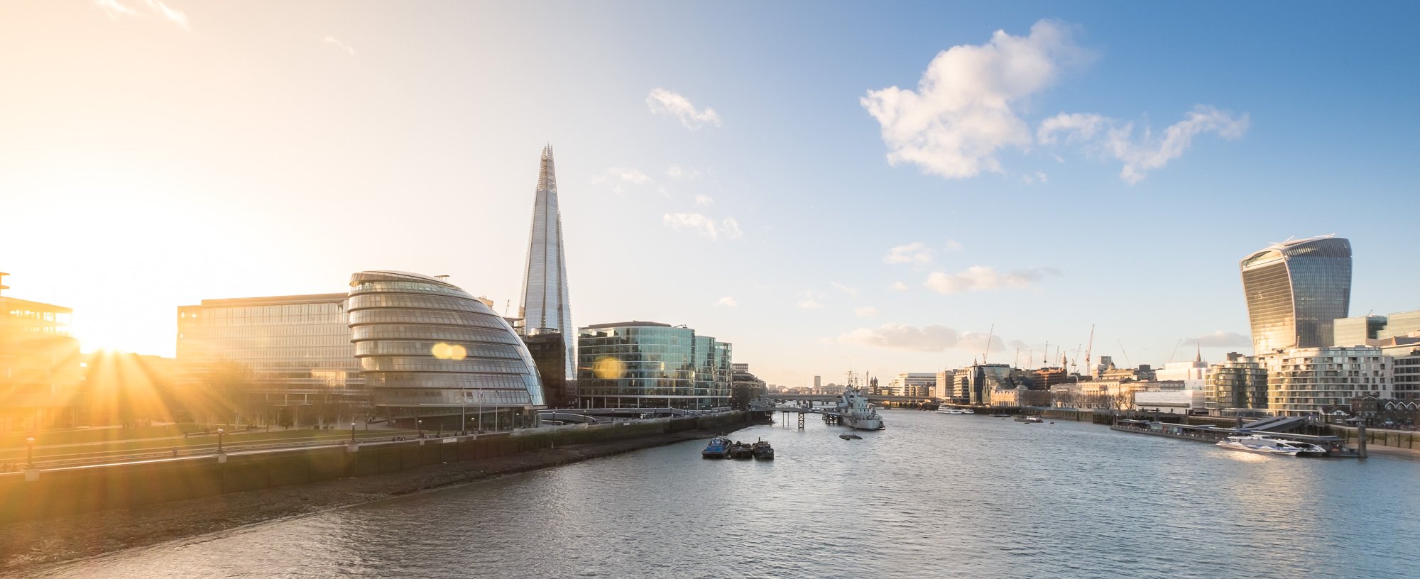 Sunset over a London city skyline with modern buildings along a river, including a skyscraper, with boats on the water and a clear sky with scattered clouds.