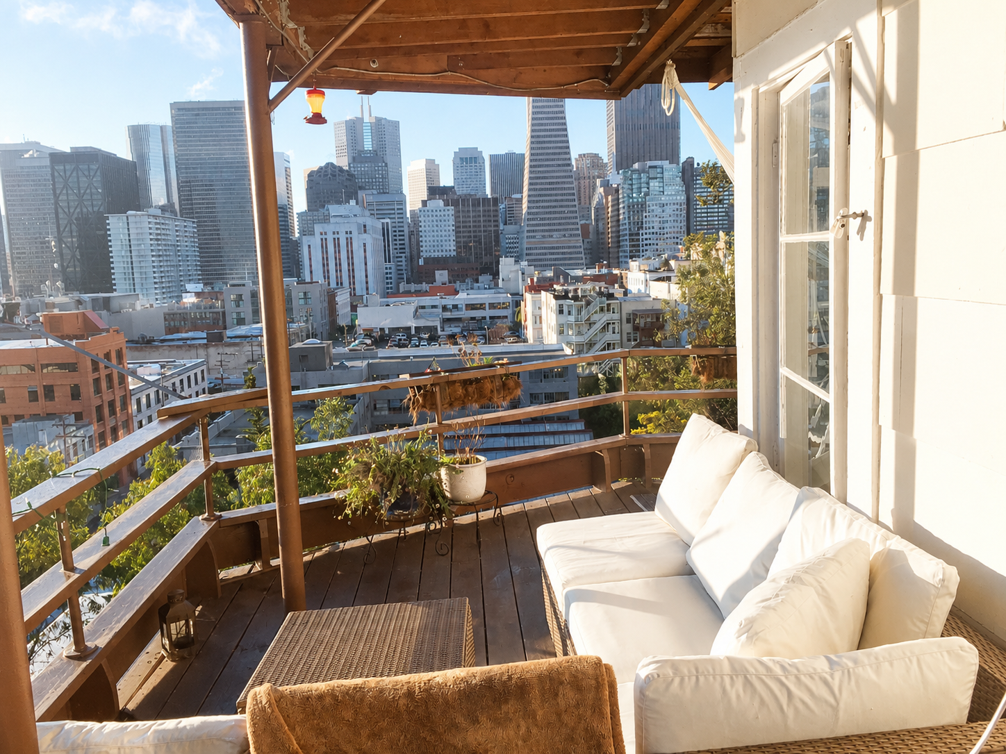 Balcony with a white cushioned sofa, potted plants, and a view of city buildings and skyscrapers in sunlight.