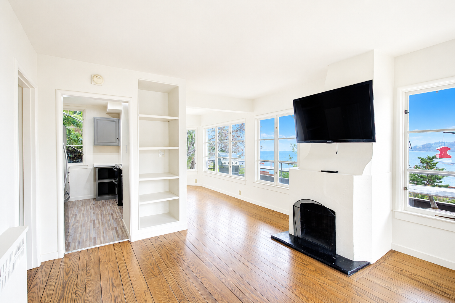 Bright living room with large windows, a wall-mounted TV, a white fireplace, and wooden floors, overlooking a view of the ocean.