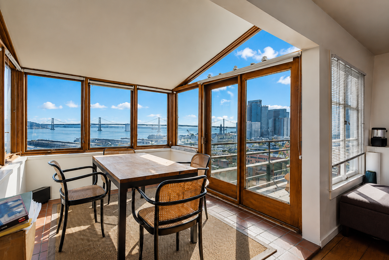 Sunlit dining area with large windows showing a view of a bridge, water, and city skyline.