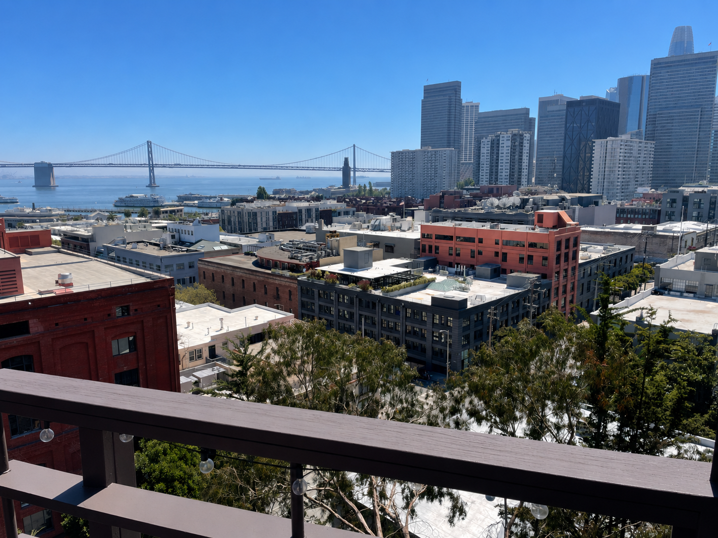 View of a city skyline with high-rise buildings, a bridge over the water, and a mix of low-rise and mid-rise buildings in the foreground, taken from a balcony with trees and a railing.