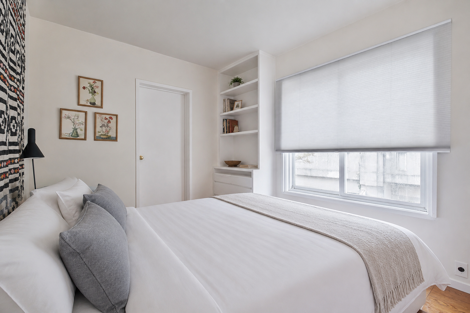 A minimalist bedroom with white walls, a white bed with grey pillows, a beige throw blanket, a black wall-mounted lamp, framed floral artwork, a built-in shelf with books and decor, and a large window with opacity blinds.