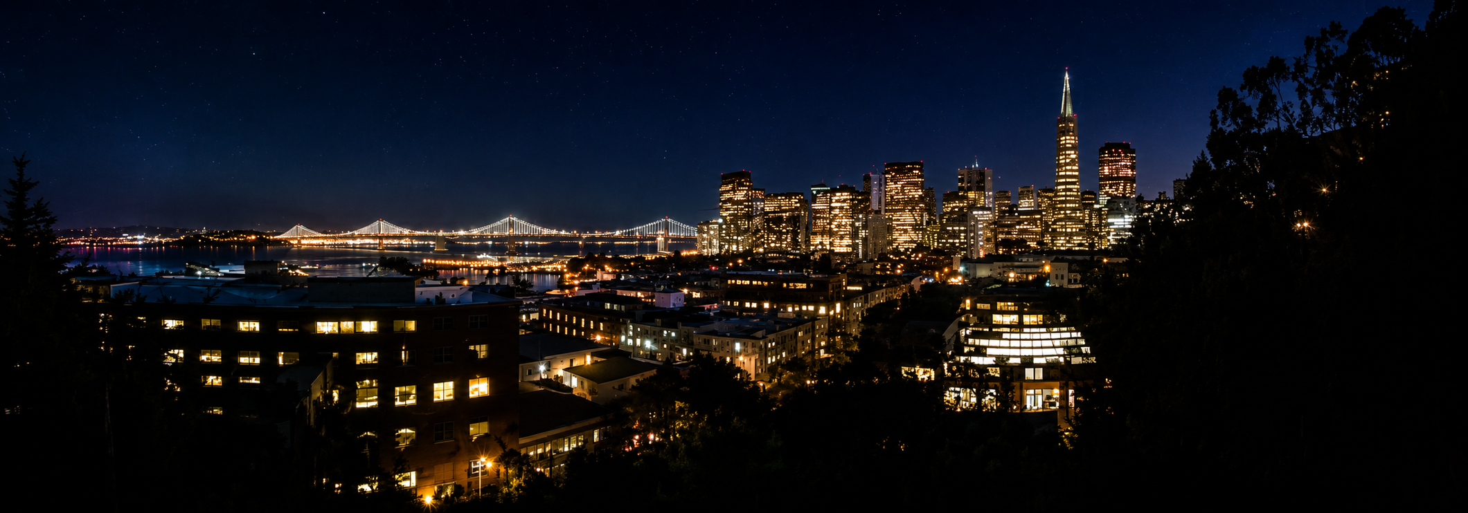 Nighttime view of San Francisco skyline with illuminated buildings, the Transamerica Pyramid, and the San Francisco-Oakland Bay Bridge in the background.