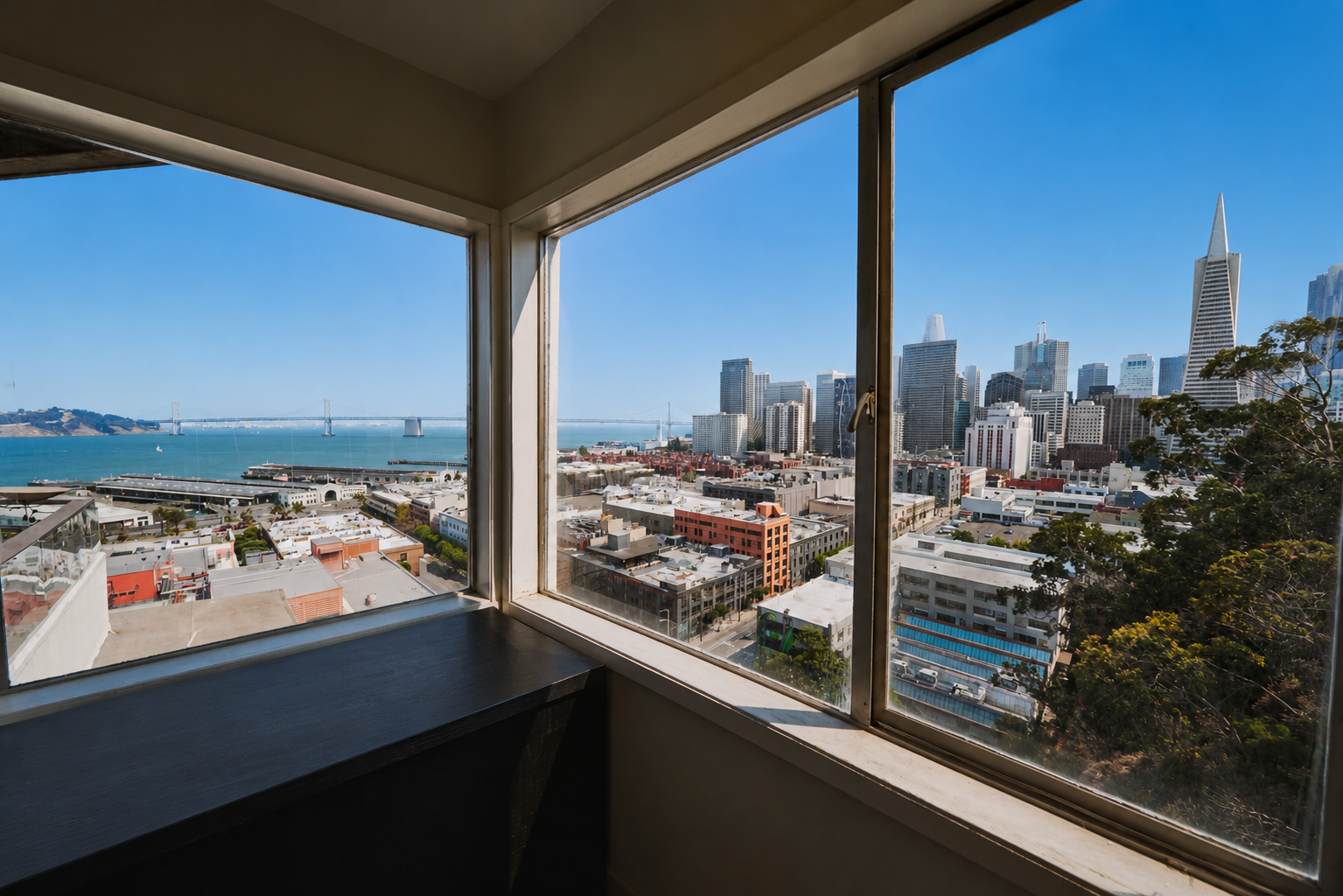 View from a high-rise window showing San Francisco skyline with the Transamerica Pyramid, the waterfront, and the Bay Bridge under a clear blue sky.