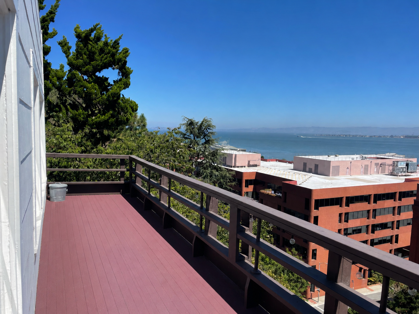 View from a balcony overlooking a city with trees, buildings, and a body of water in the distance under a clear blue sky.