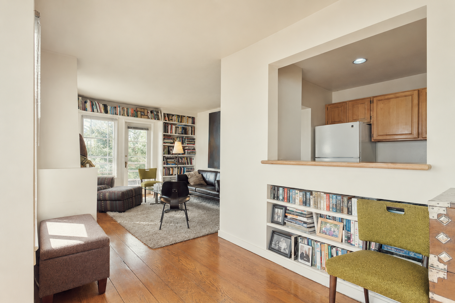 Living room with wooden floors, a bookcase filled with books, a black leather sofa, a green armchair, a patterned ottoman, and large windows with a door leading outside.