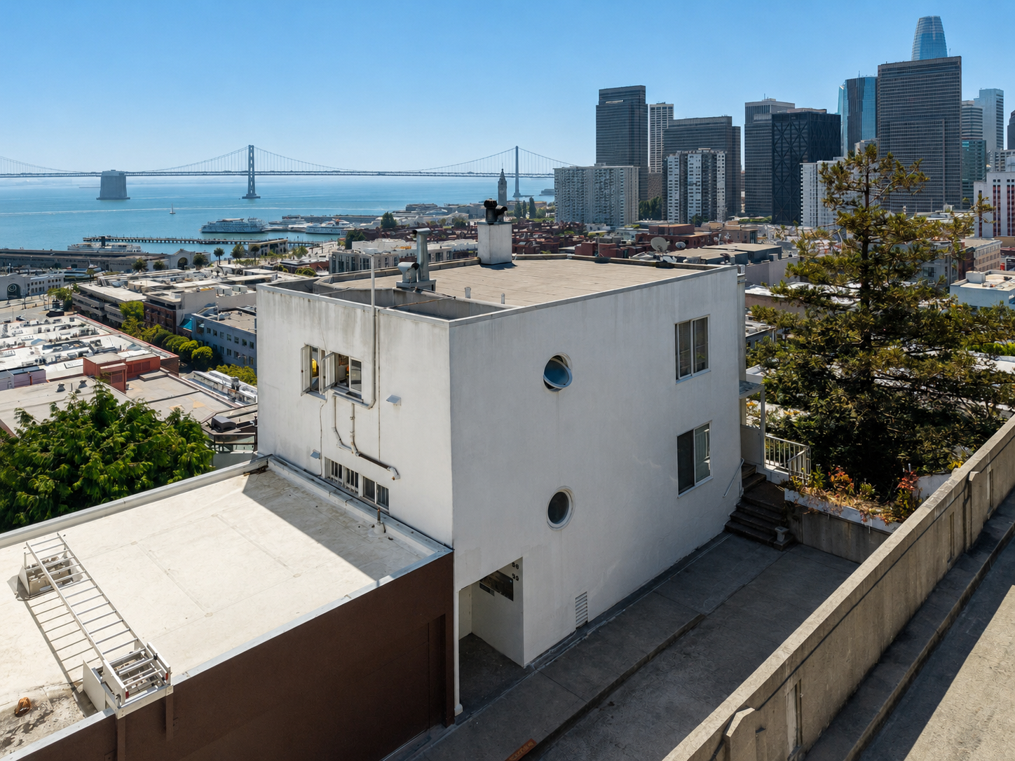 View of San Francisco skyline, including buildings, Alcatraz Island, bridge, and bay with a white residential building and greenery in foreground.