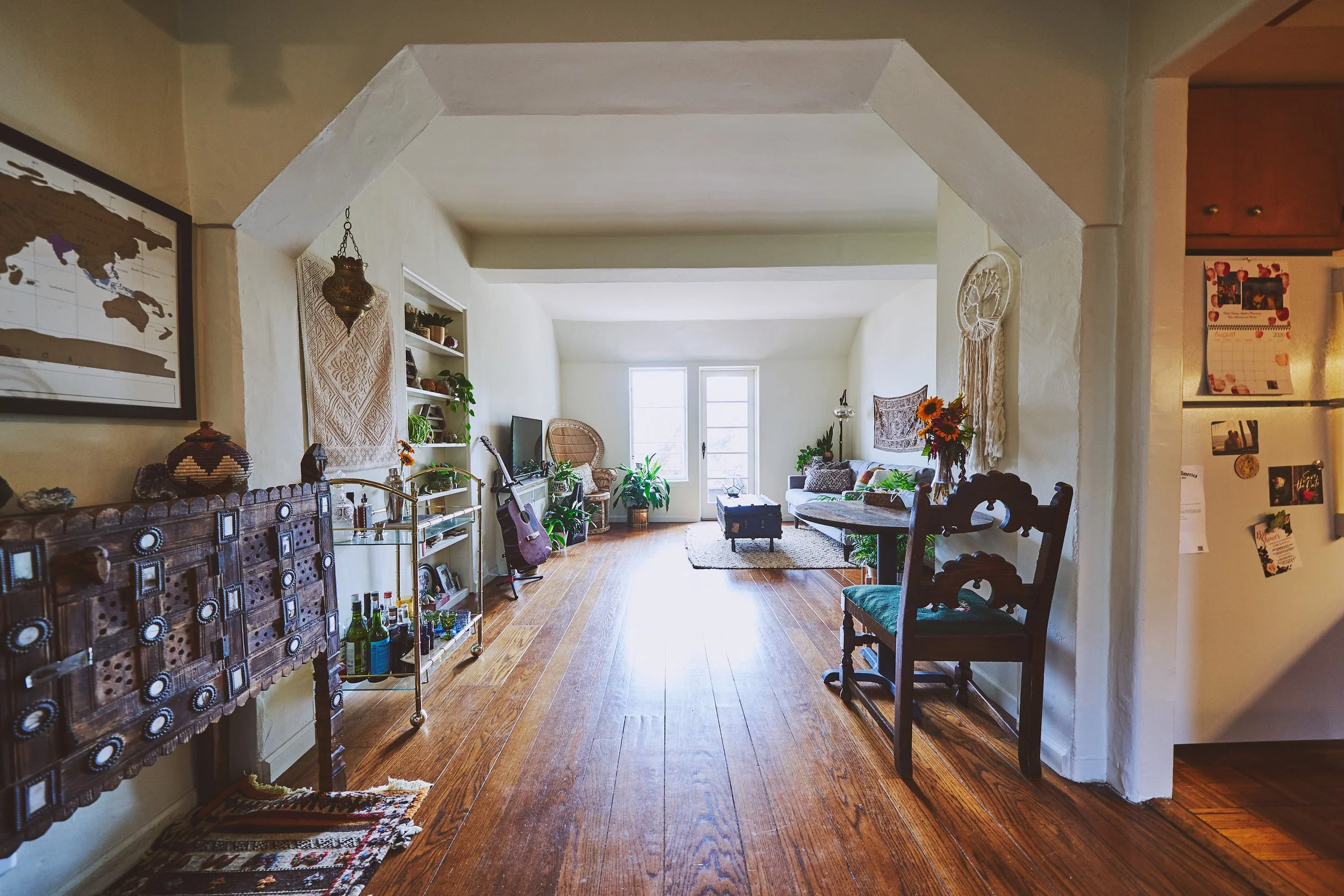 View of a cozy living room with wooden floors, white walls, large windows, a white sofa, a round wooden table, and various plants and decorations, including guitars and framed pictures.